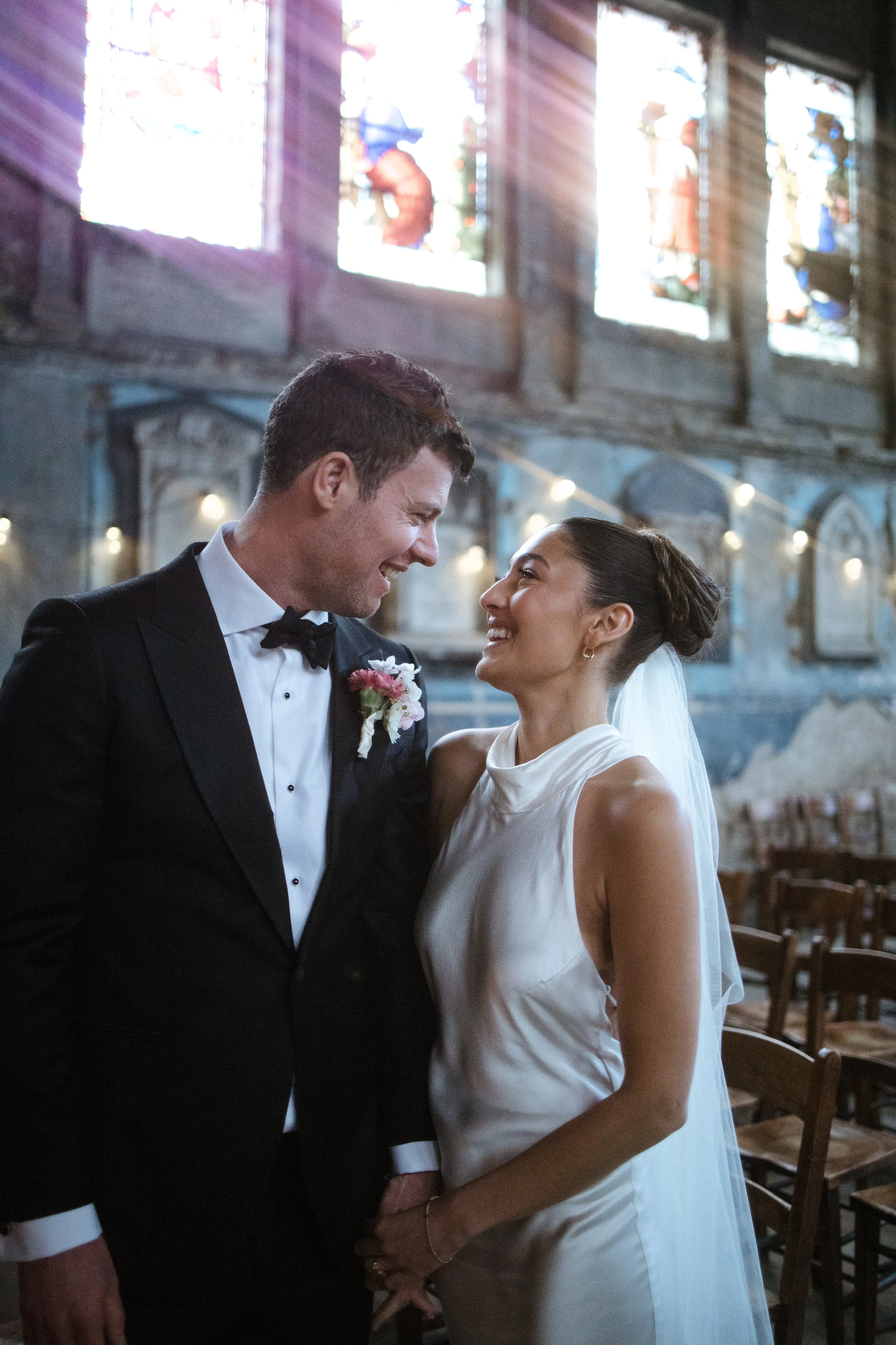 A happy couple, dressed as a groom and bride, sharing a joyful moment at their wedding reception in a rustic indoor venue with stained glass windows and string lights.