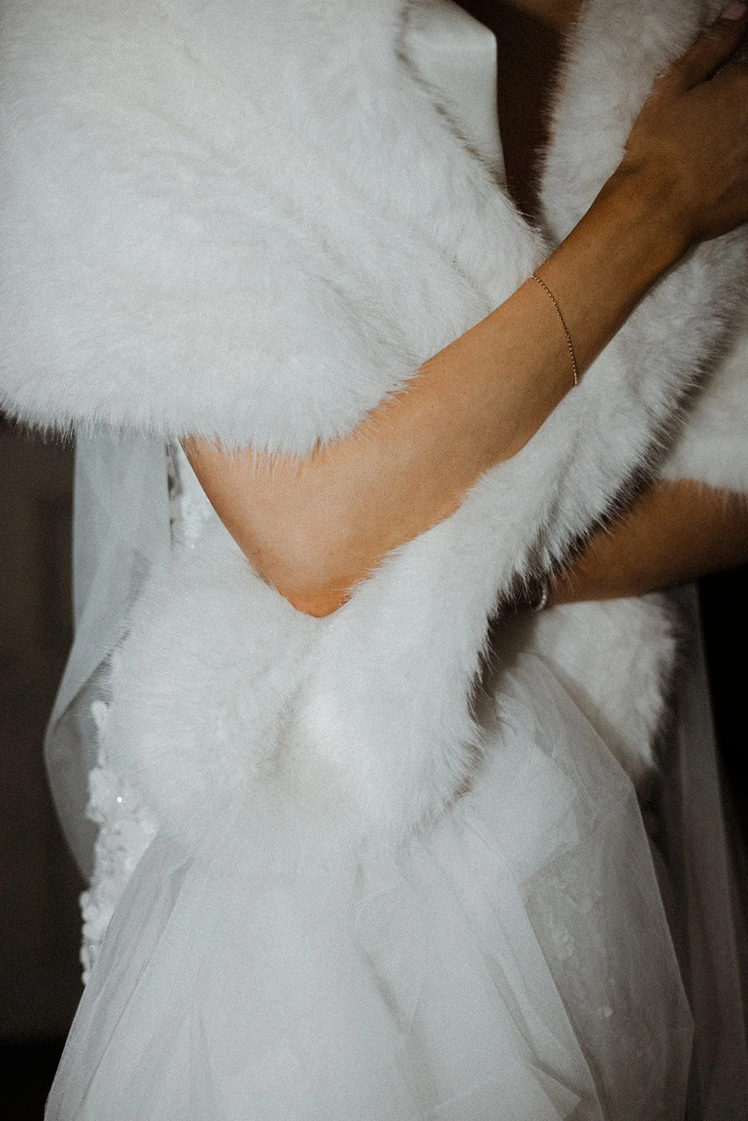 Close-up of a person wearing a white fur coat and a delicate gold bracelet, with a white dress visible underneath.