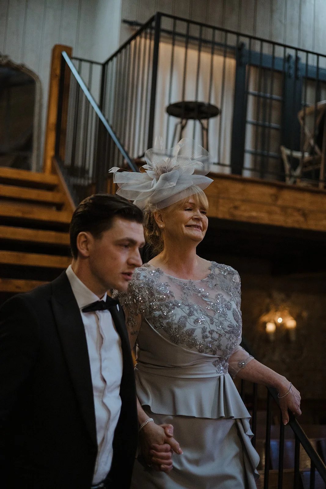 A woman wearing an elaborate hat and dress holding a young man's hand during a formal event in a rustic venue.