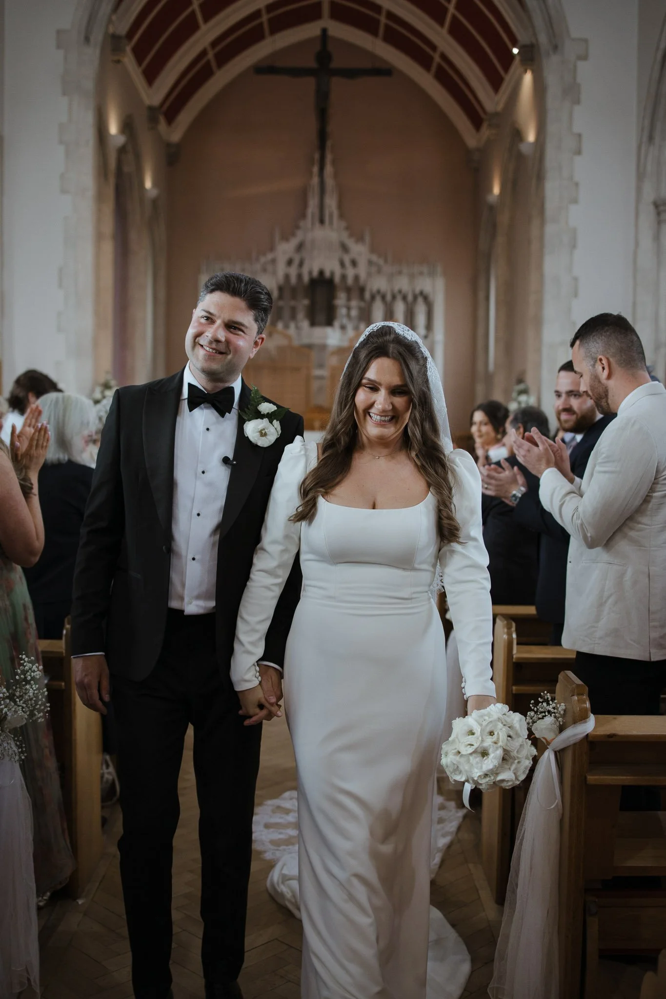 A bride and groom walking down the aisle in a church, holding hands, smiling, surrounded by applauding guests.