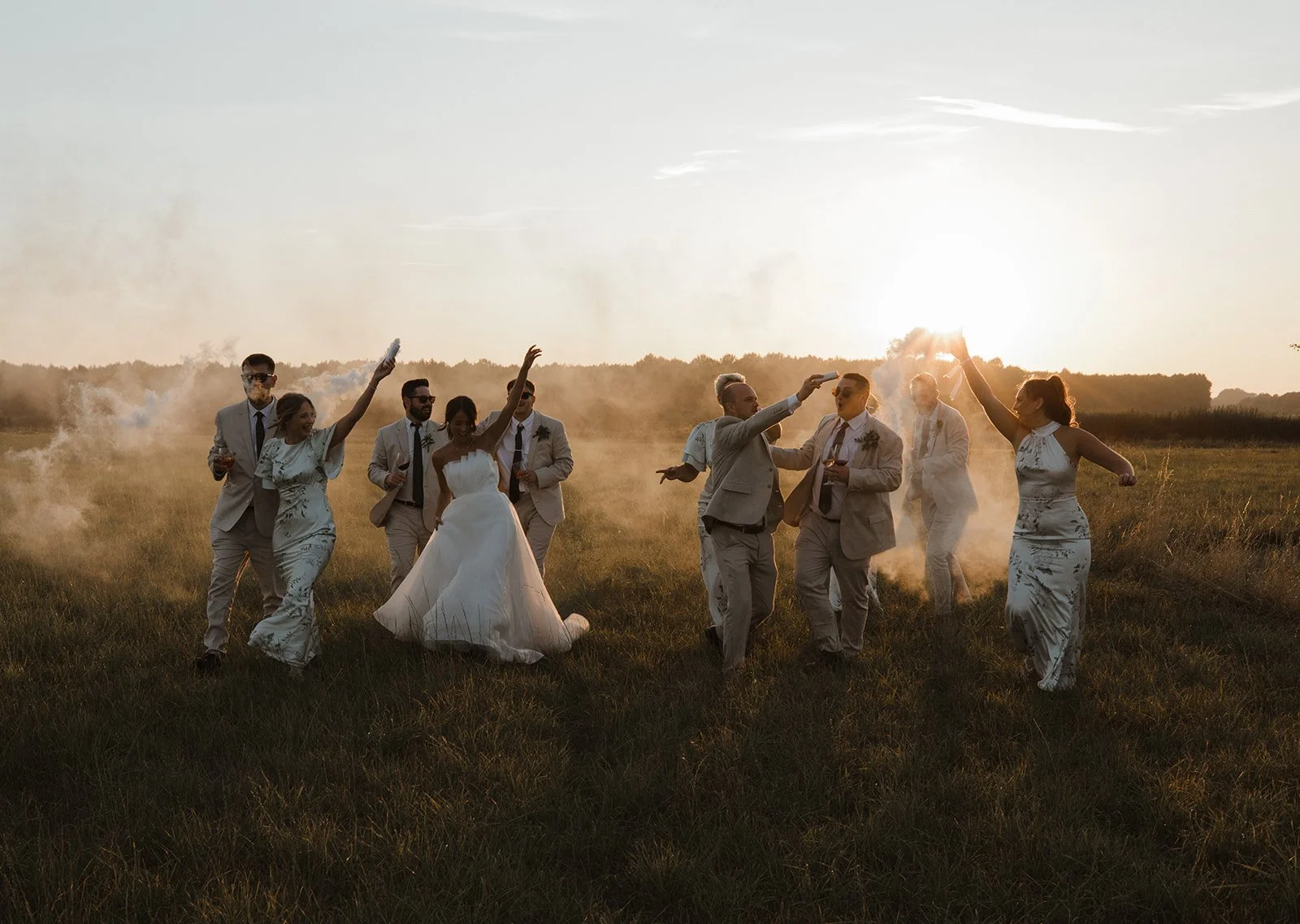 Group of people celebrating outdoors at sunset, with smoke and sun rays in the background, dressed in wedding attire and enjoying drinks.
