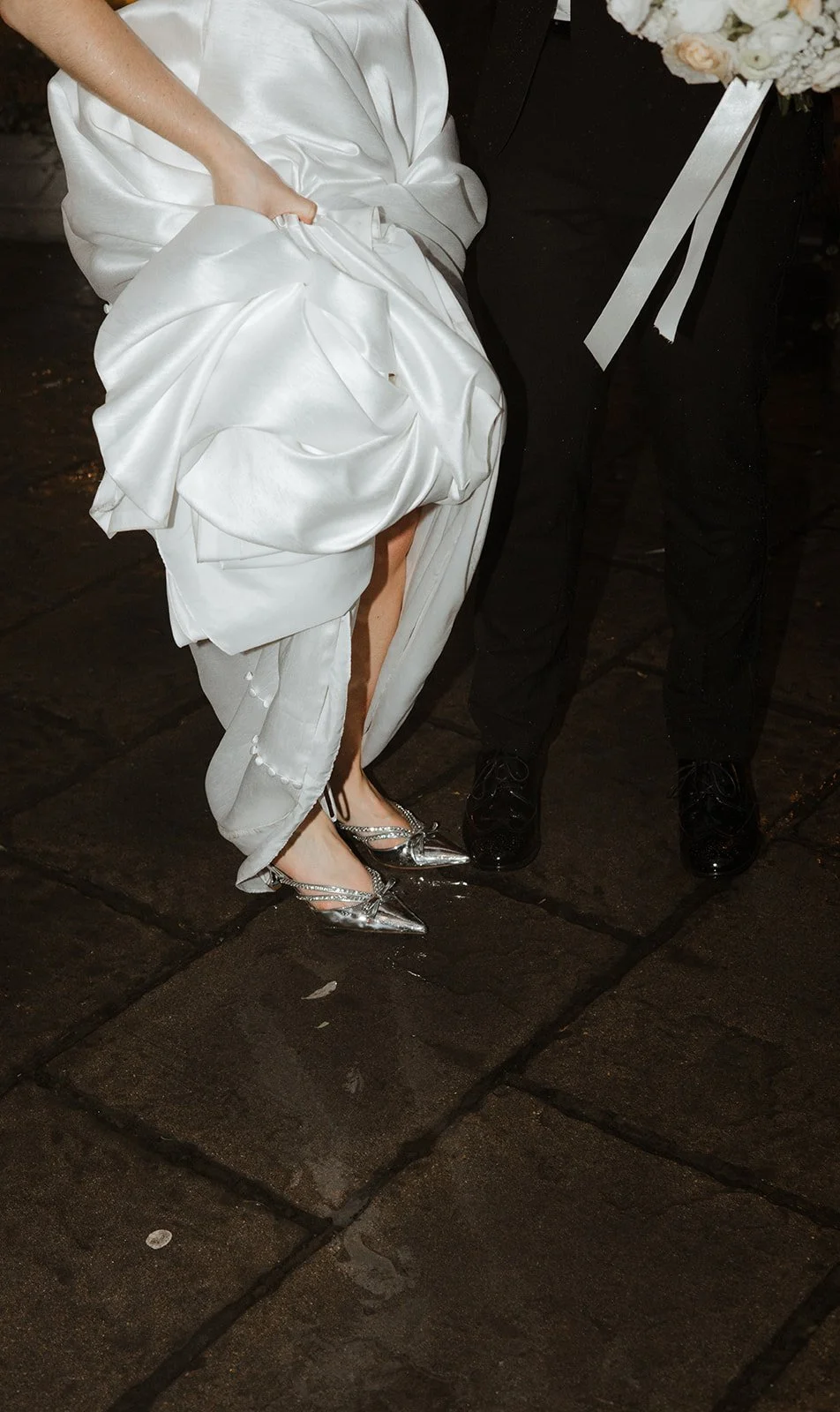 Close-up of a bride in a white wedding gown lifting the skirt to show her silver high heels, standing next to a groom in black shoes, on dark stone flooring.