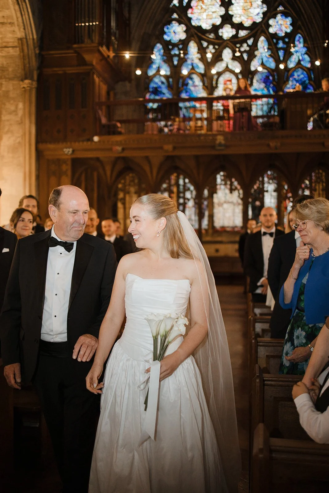 A bride and her father walk down the aisle of a church, surrounded by wedding guests, with stained glass windows and wooden architecture in the background.