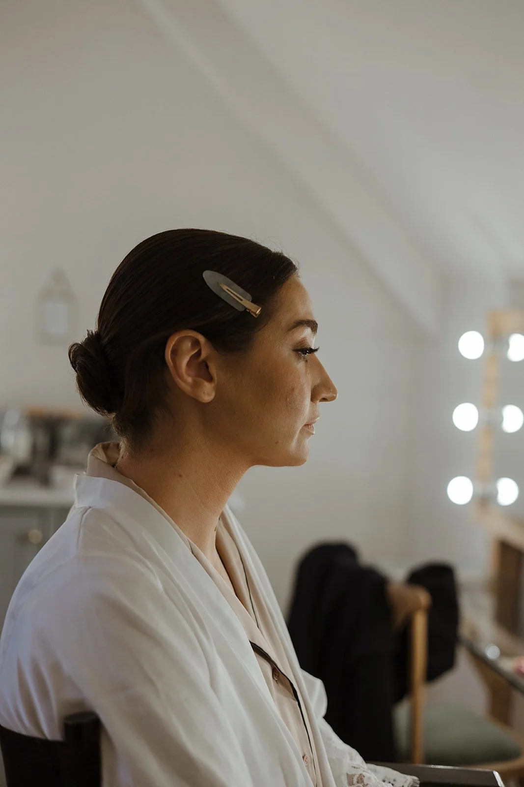 A woman with dark hair in a low bun with a hair clip, wearing a white robe, sitting in front of mirrors with bright lights, possibly getting ready.