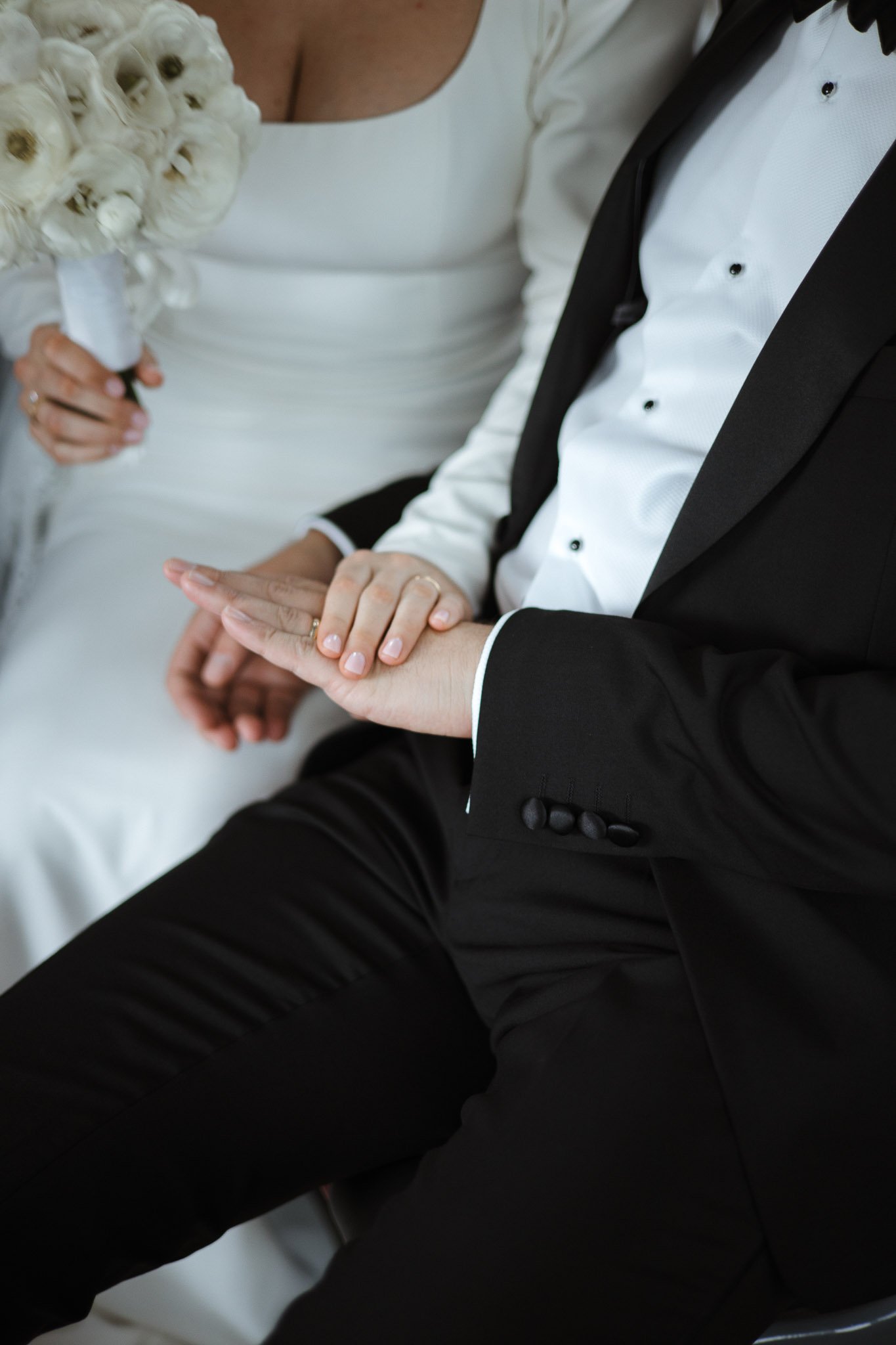 A bride and groom sitting together, with the bride holding a bouquet of white flowers, the groom wearing a black tuxedo, and their hands resting on each other's.