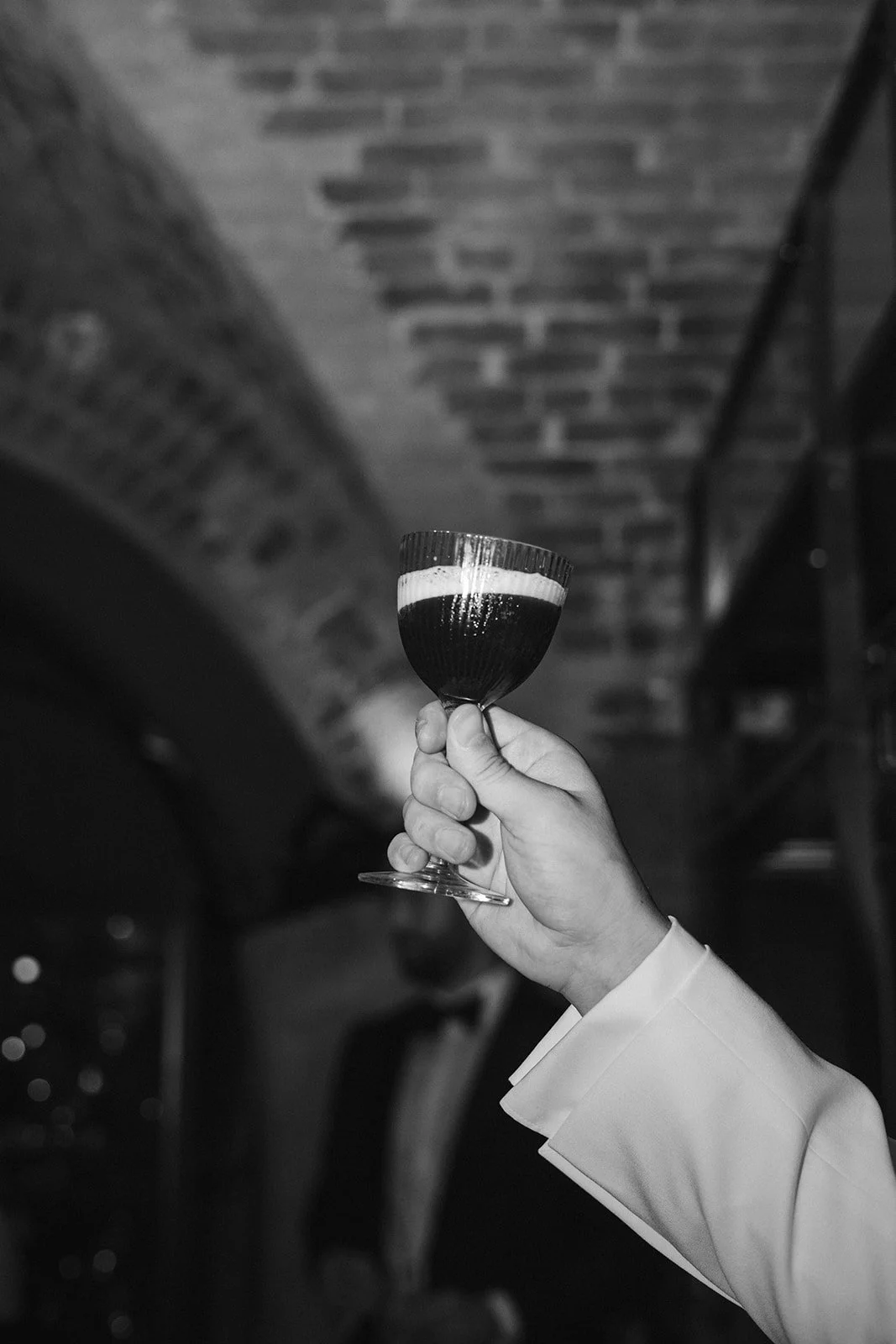 A person holding a cocktail glass filled with layered dark and light drink, with a background of a brick ceiling and warm lighting.