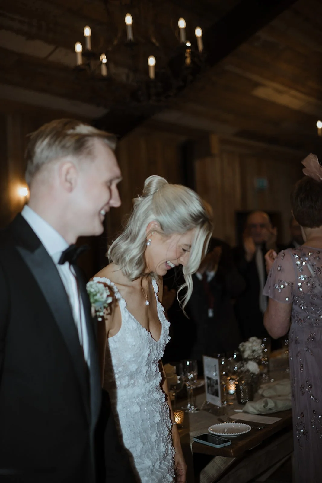 A bride and groom smiling and standing side by side at a wedding reception, with other guests in the background, inside a wooden venue with candlelit chandeliers.