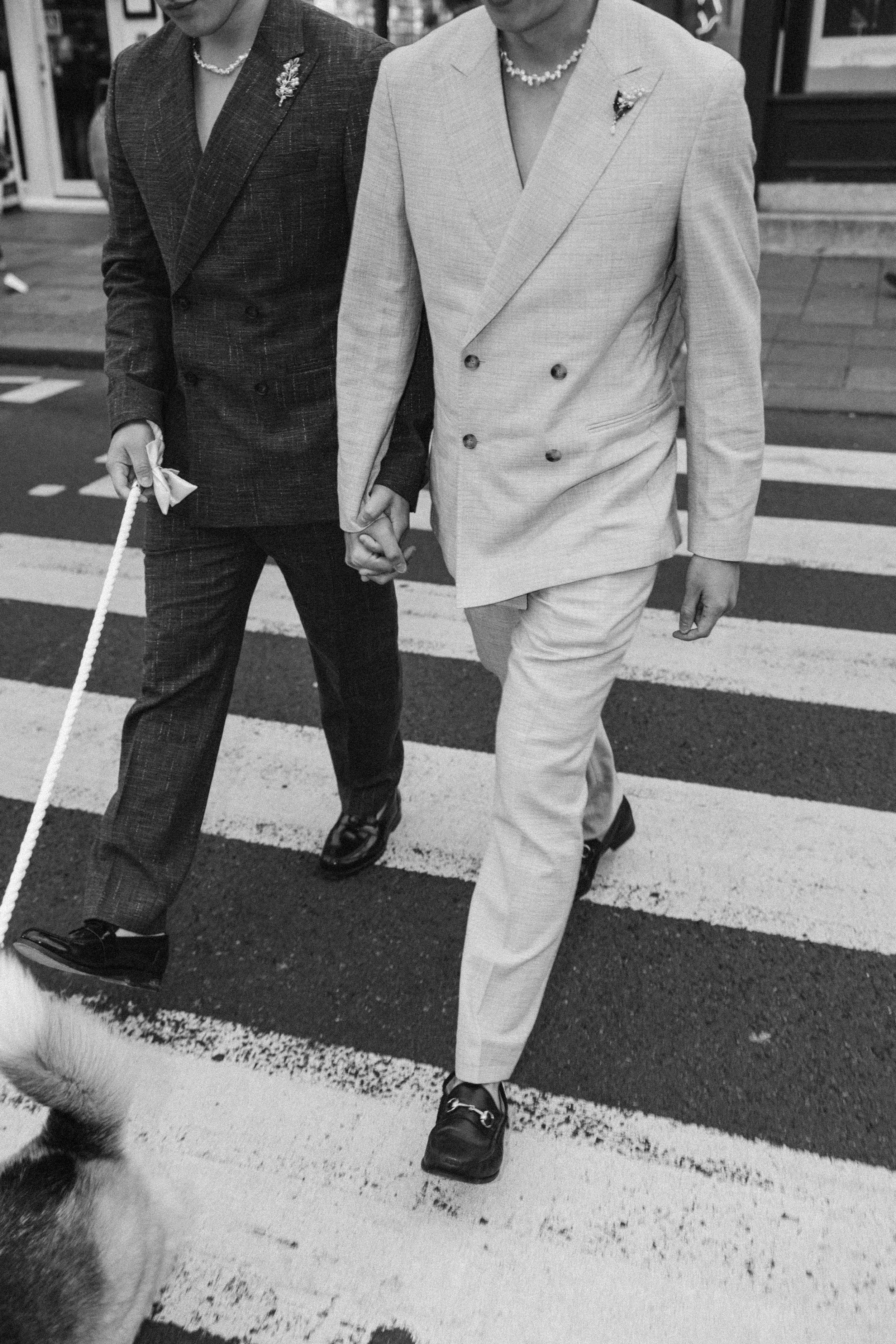Two people holding hands and walking on a crosswalk, dressed in formal suits. One person is wearing a dark plaid suit, and the other person is wearing a light-colored double-breasted suit. Both are wearing necklaces and brooches.