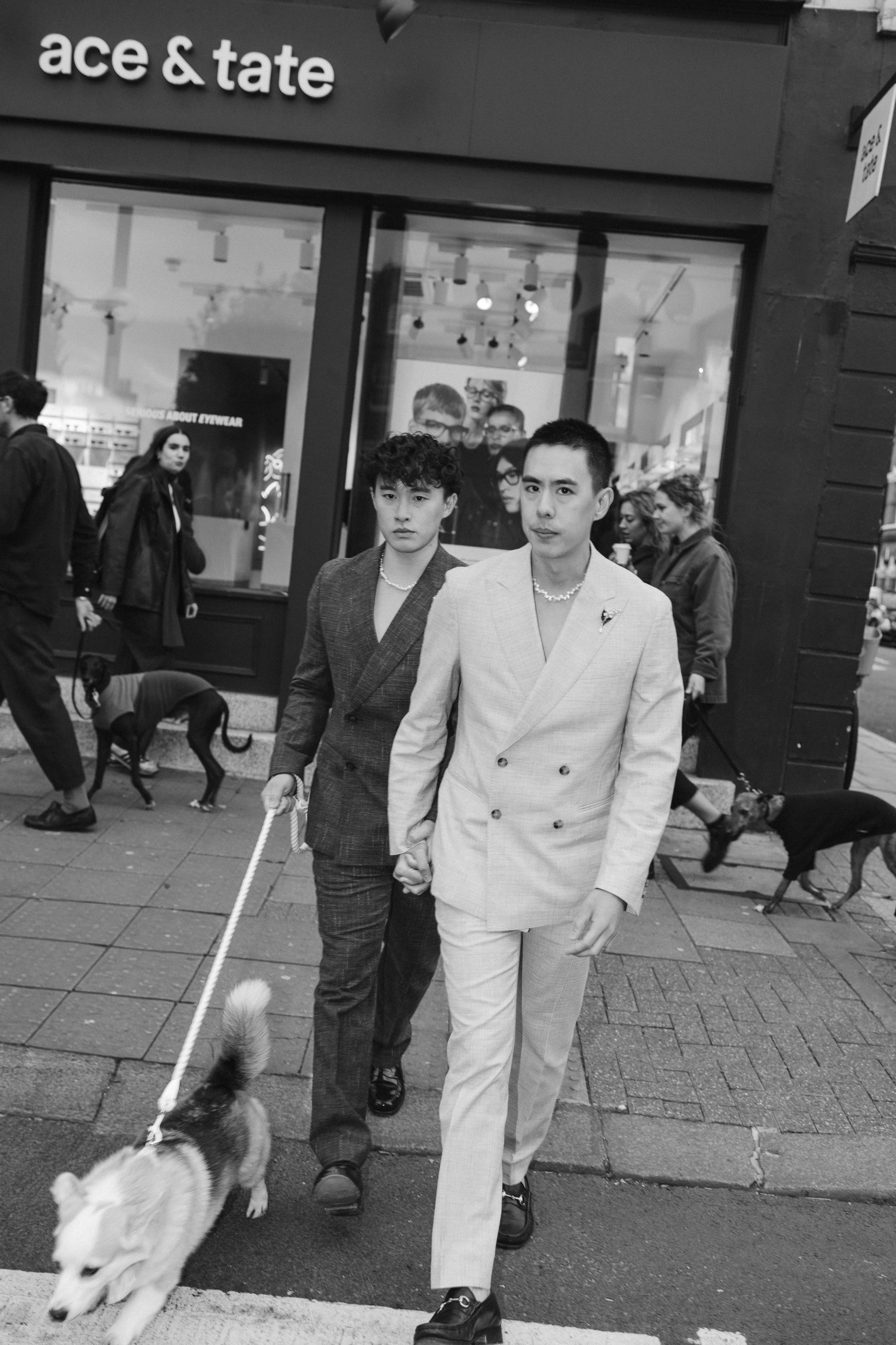 Two men dressed in stylish suits walking down a city street holding hands, with dogs on leashes. People converse and walk behind them, with a storefront sign visible above reading "ace & tate" and posters inside