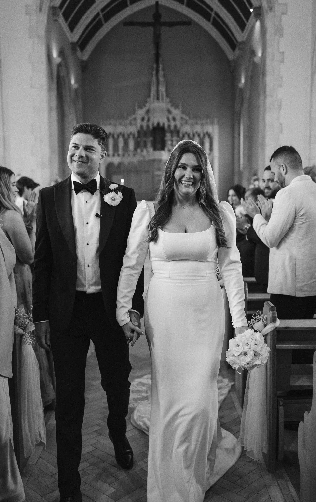 Black and white photo of a bride and groom walking down the aisle in a church, holding hands, surrounded by wedding guests.