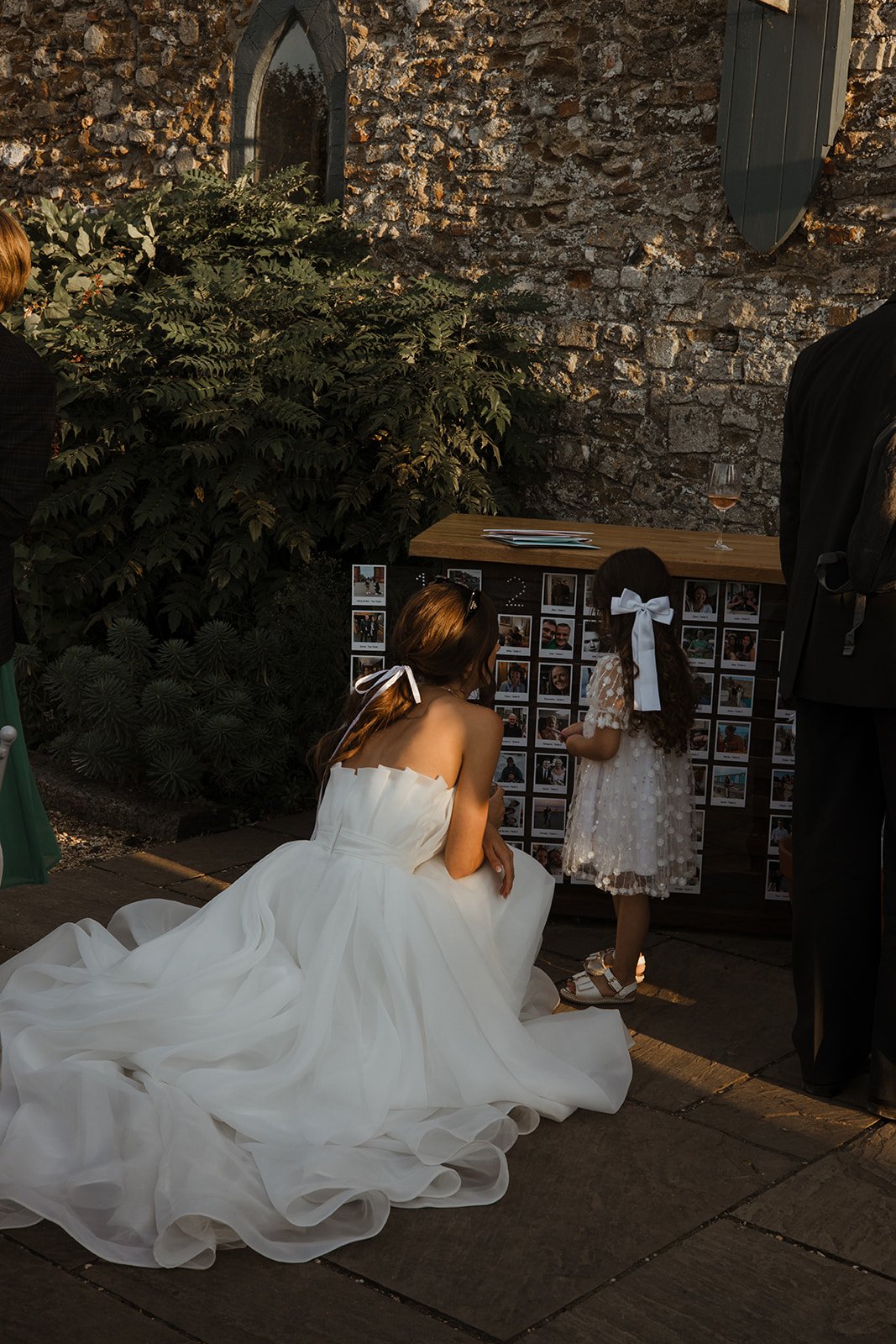 A woman in a white wedding dress kneeling and talking to a young girl in a white dress with a large white bow, outdoors near a stone wall and greenery, with a glass of wine on a wooden table nearby.