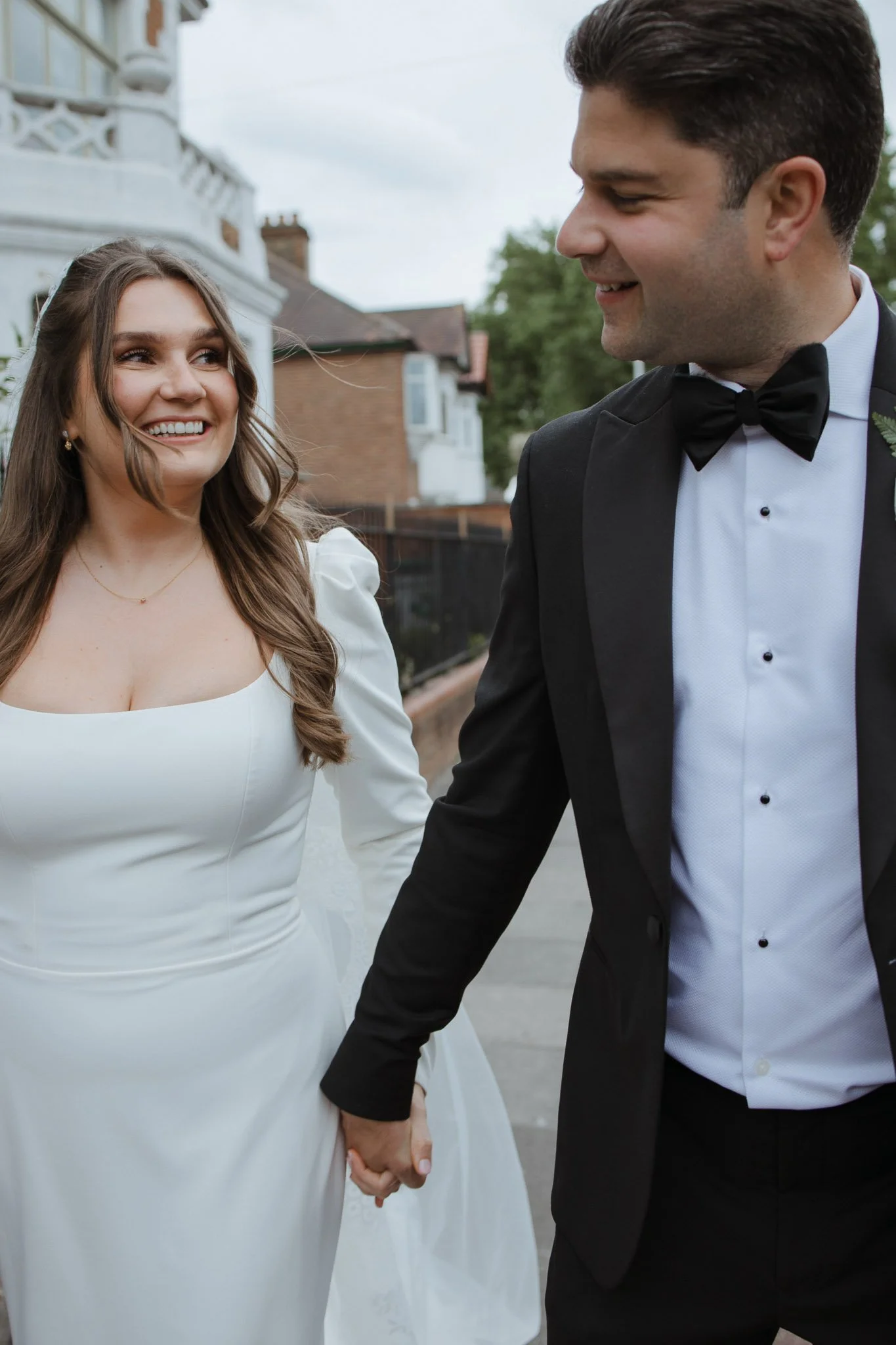 A smiling bride and groom holding hands outdoors, dressed in wedding attire.
