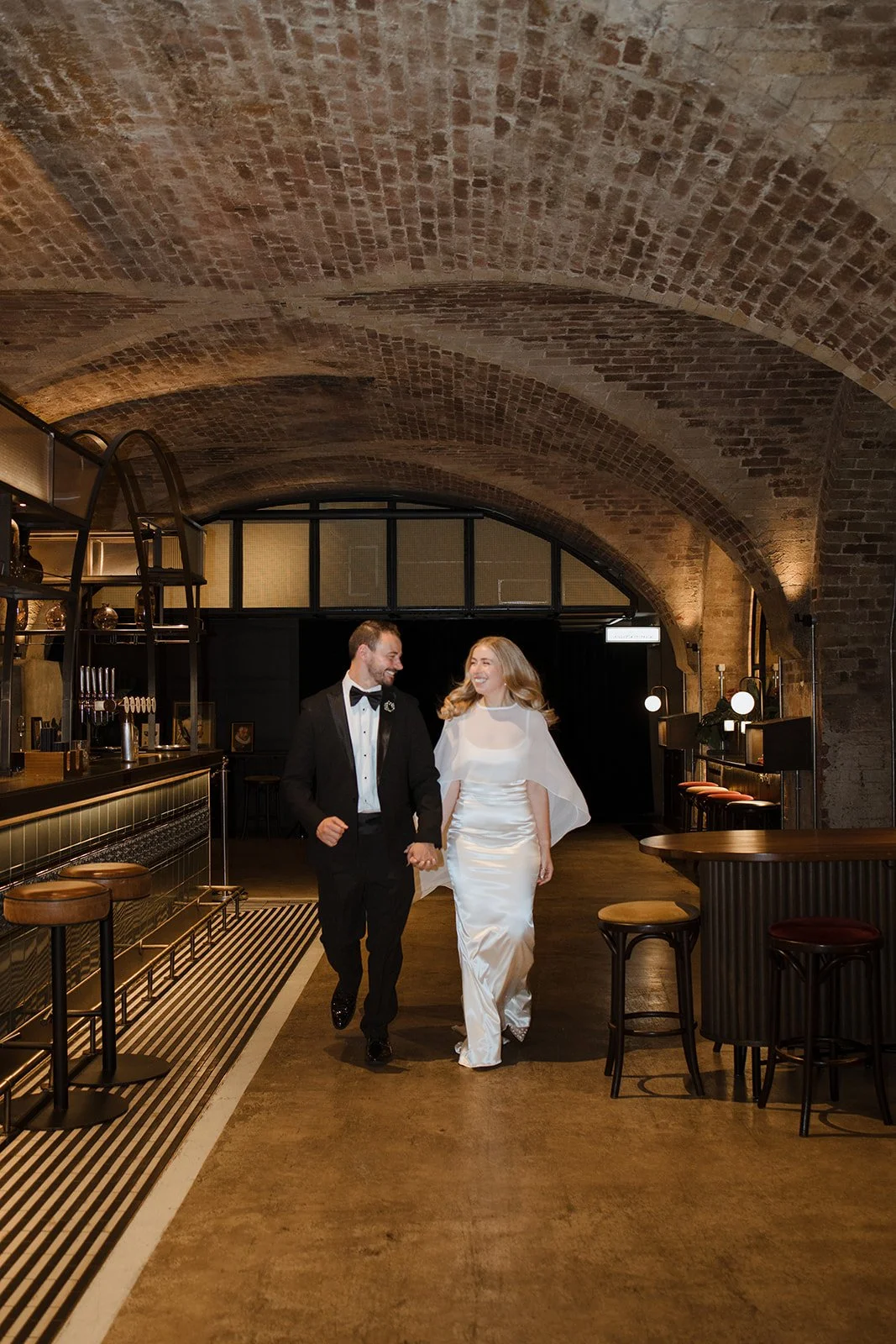 A newlywed couple holding hands and smiling as they walk through a stylish, dimly lit bar with exposed brick arches and contemporary decor.