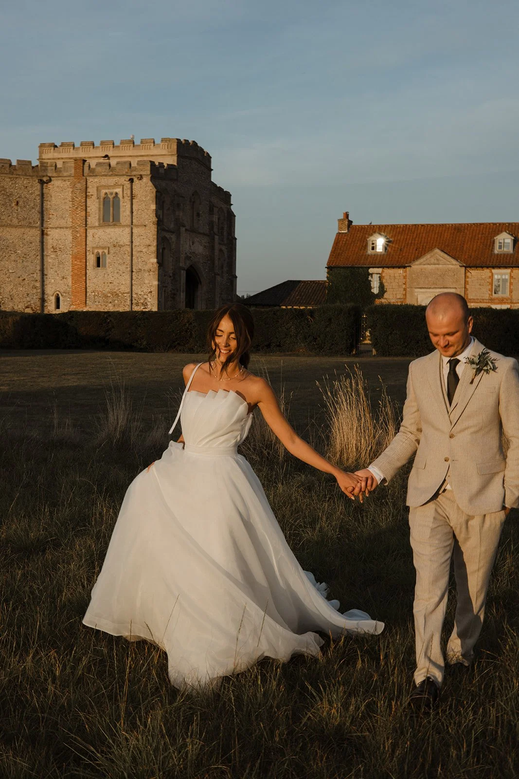 Bride and groom holding hands walking through a grassy field at sunset, with an old castle and traditional buildings in the background.