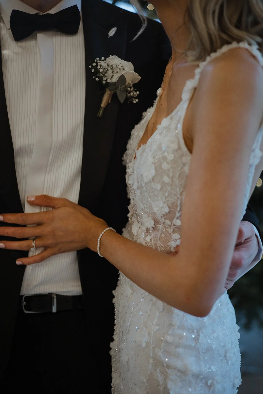 Close-up of a bride and groom during their wedding ceremony, showing the bride in a white lace wedding dress and the groom in a black tuxedo with a bowtie, with the bride's hand and wedding ring visible.