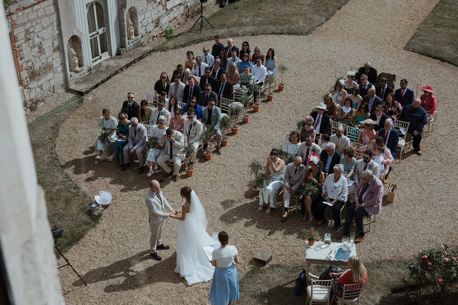 Outdoor wedding ceremony with guests seated in rows, bride and groom exchanging vows, officiant present, and guests wearing formal attire in a stone courtyard.