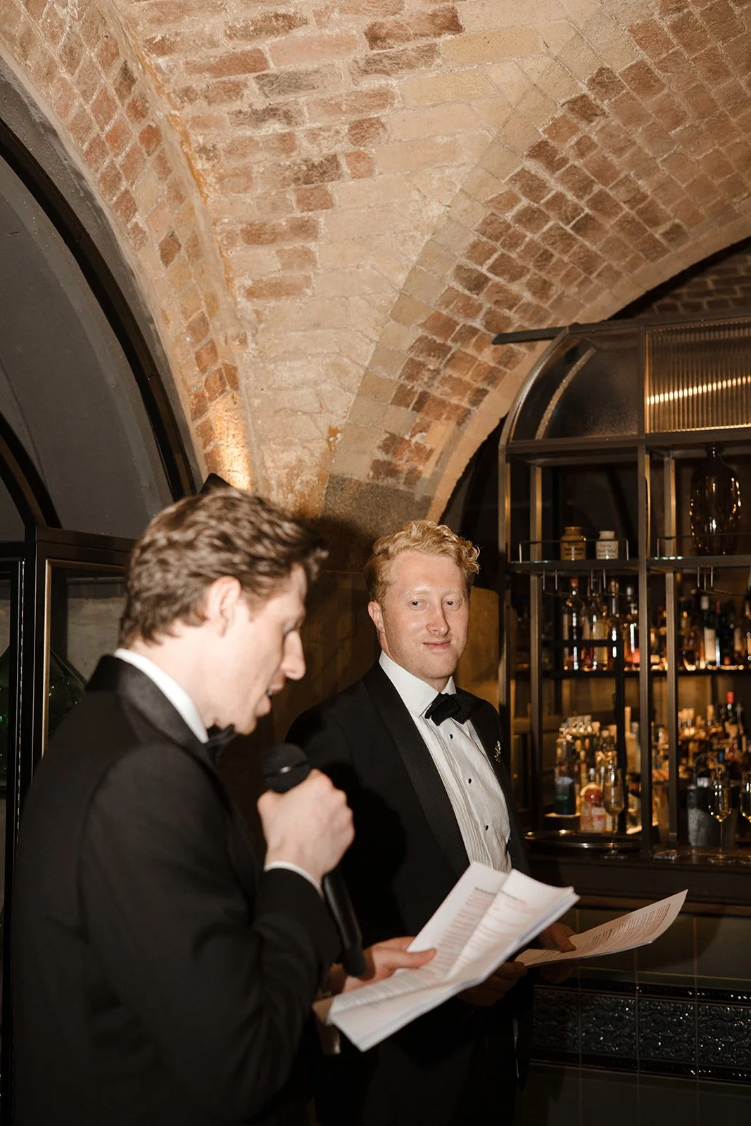 Two men in tuxedos, one reading from a paper, the other looking at the camera, in a dimly lit bar or restaurant with exposed brick ceiling and liquor shelves.