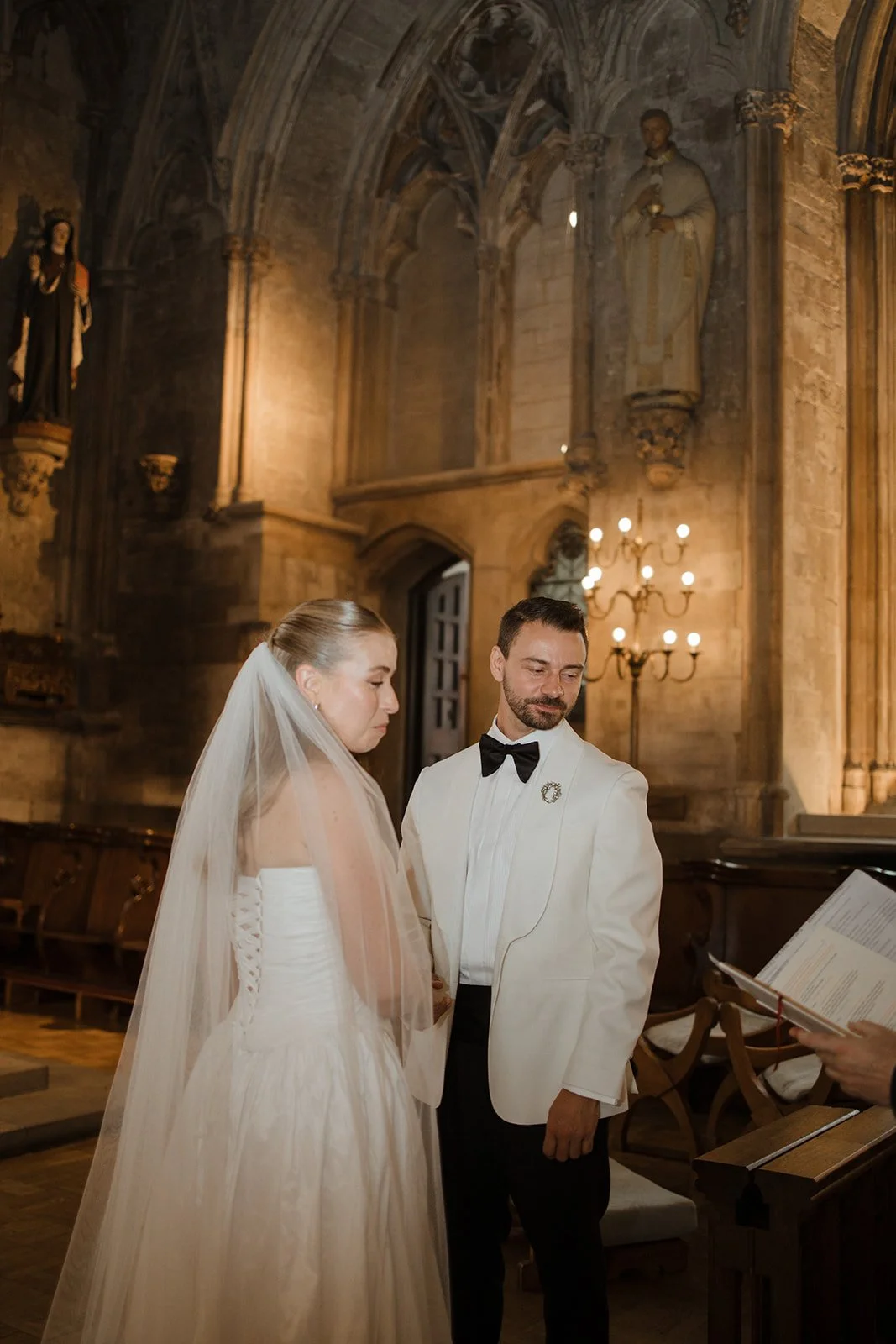 A bride and groom exchanging vows at a wedding ceremony inside a historic church with stone walls and religious statues, with a person reading from a book.