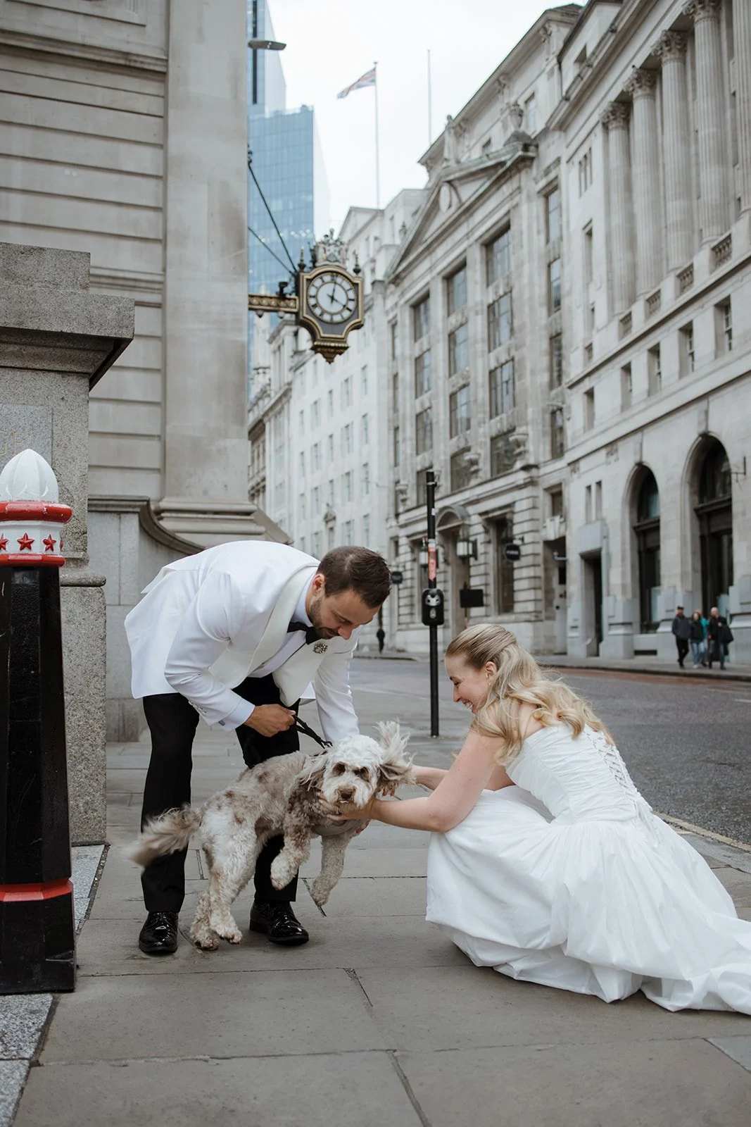 A man in a tuxedo and a woman in a wedding dress are interacting with a dog on a city sidewalk. The man is smiling while holding the dog, and the woman is kneeling and petting the dog. The scene is set in an urban area with historic buildings and a c