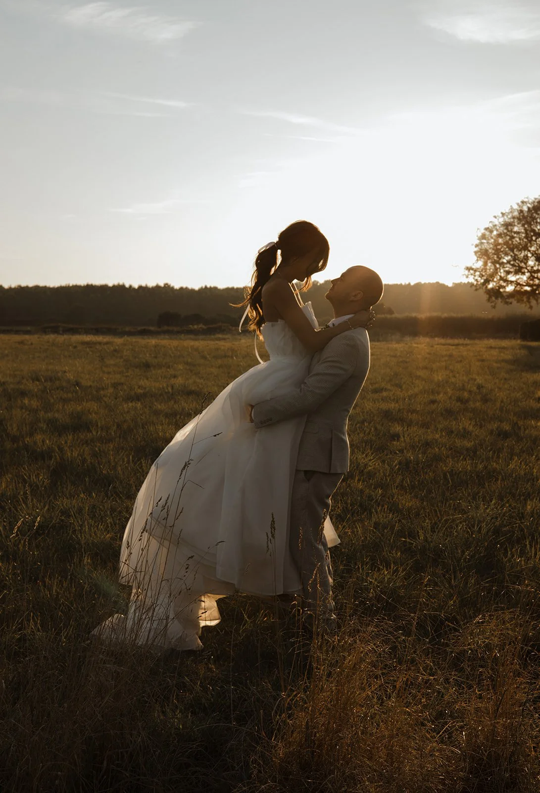 A couple dressed in wedding attire, with the man lifting the woman in a field at sunset, creating a romantic silhouette.