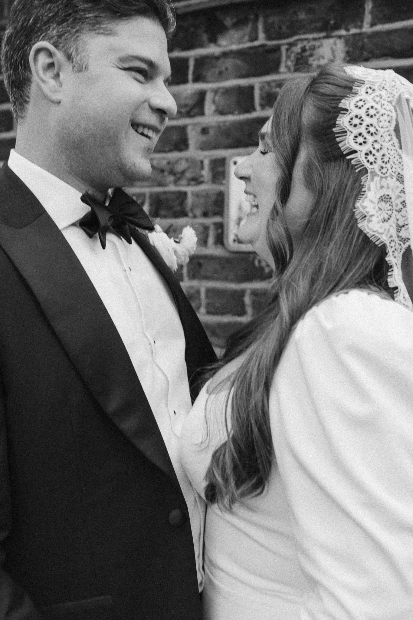 A black and white photo of a smiling couple, the groom in a tuxedo and the bride in a white dress with a lace veil, standing close together against a brick wall.