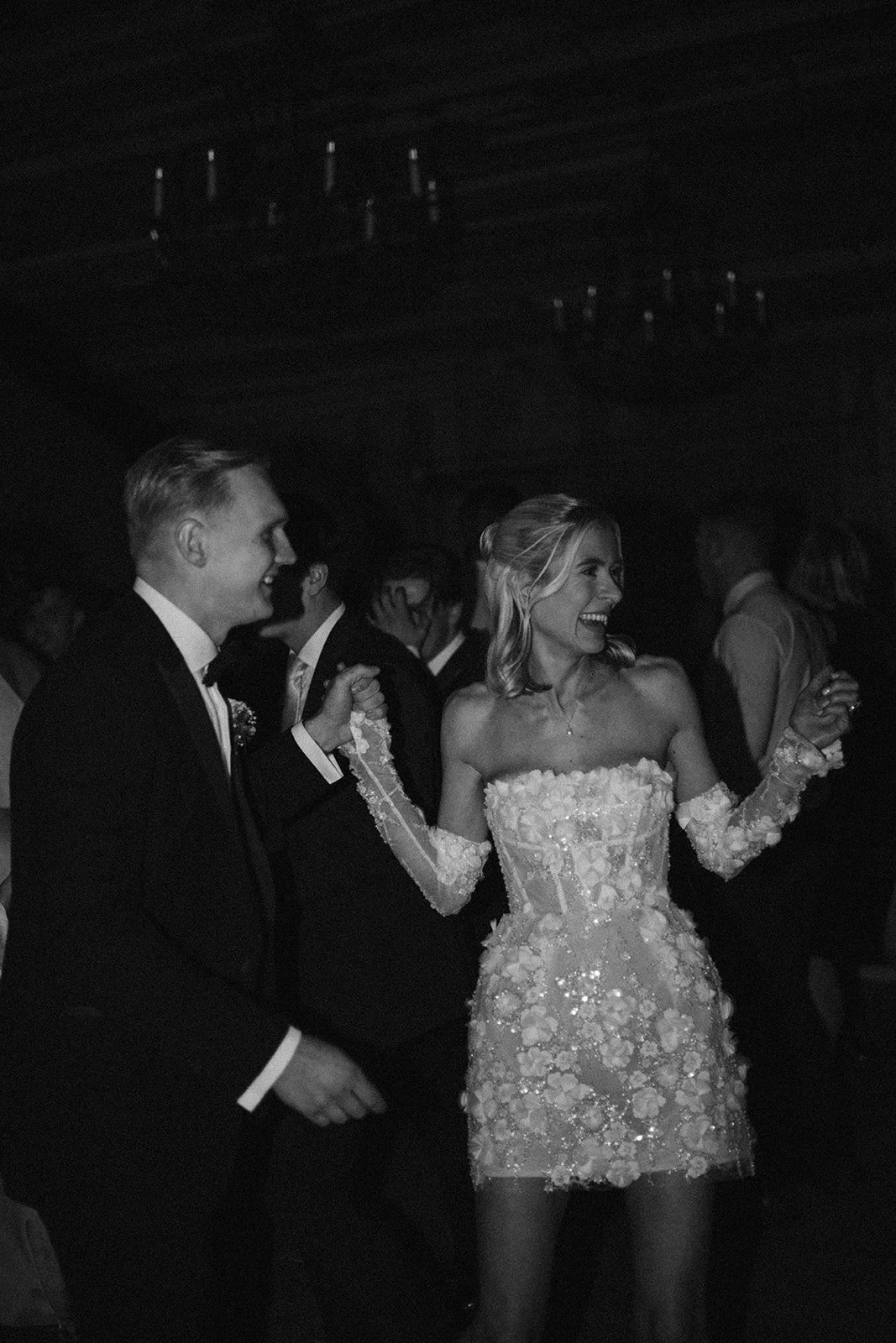 A woman in a floral, off-the-shoulder dress dancing with a man in a tuxedo at a party, both smiling and holding hands. Other people are visible in the background.