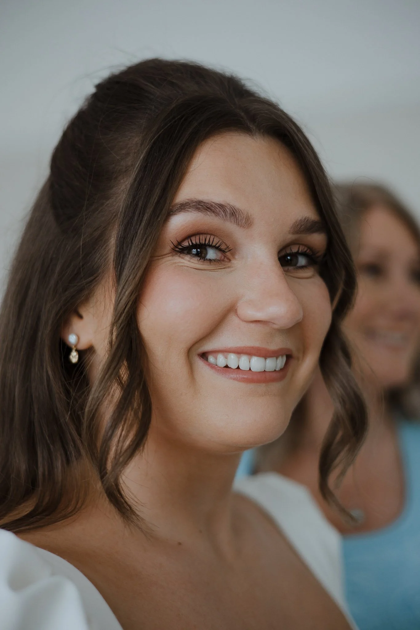 Close-up of a smiling woman with brown hair, wearing pearl earrings, and a white top, with another woman blurred in the background.