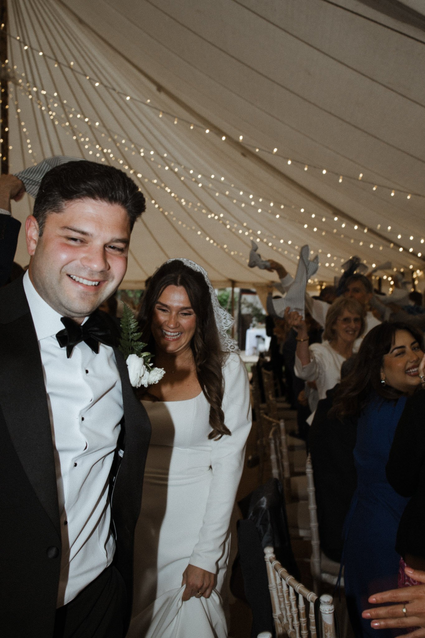 Smiling bride in white wedding dress and groom in black tuxedo at wedding reception, under a decorated tent with string lights and guests in the background.