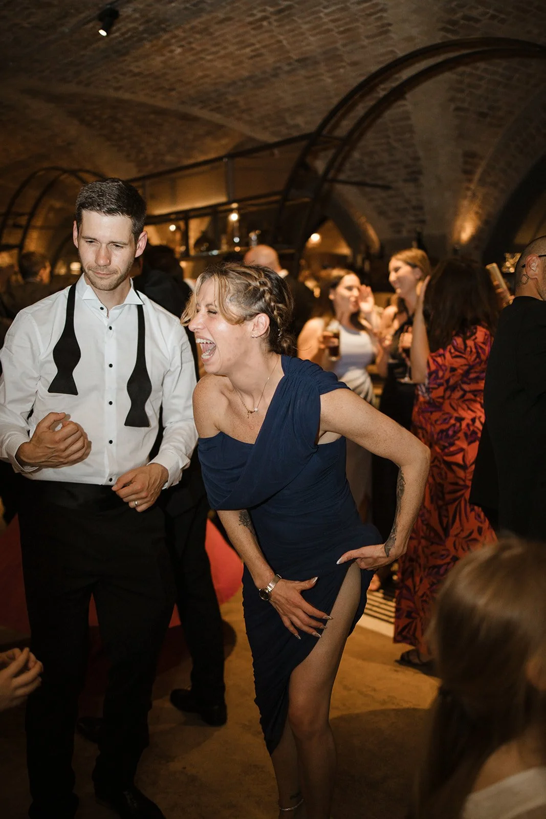 People dancing and socializing at a party or celebration in a warmly lit, arched brick venue.