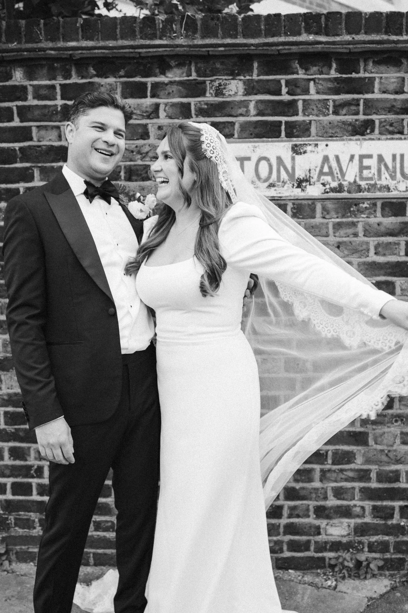 A wedding photograph of a smiling bride and groom standing in front of a brick wall with a street sign that reads "TON AVENUE." The bride is wearing a white gown and lace veil, and the groom is dressed in a black tuxedo with a bow tie.