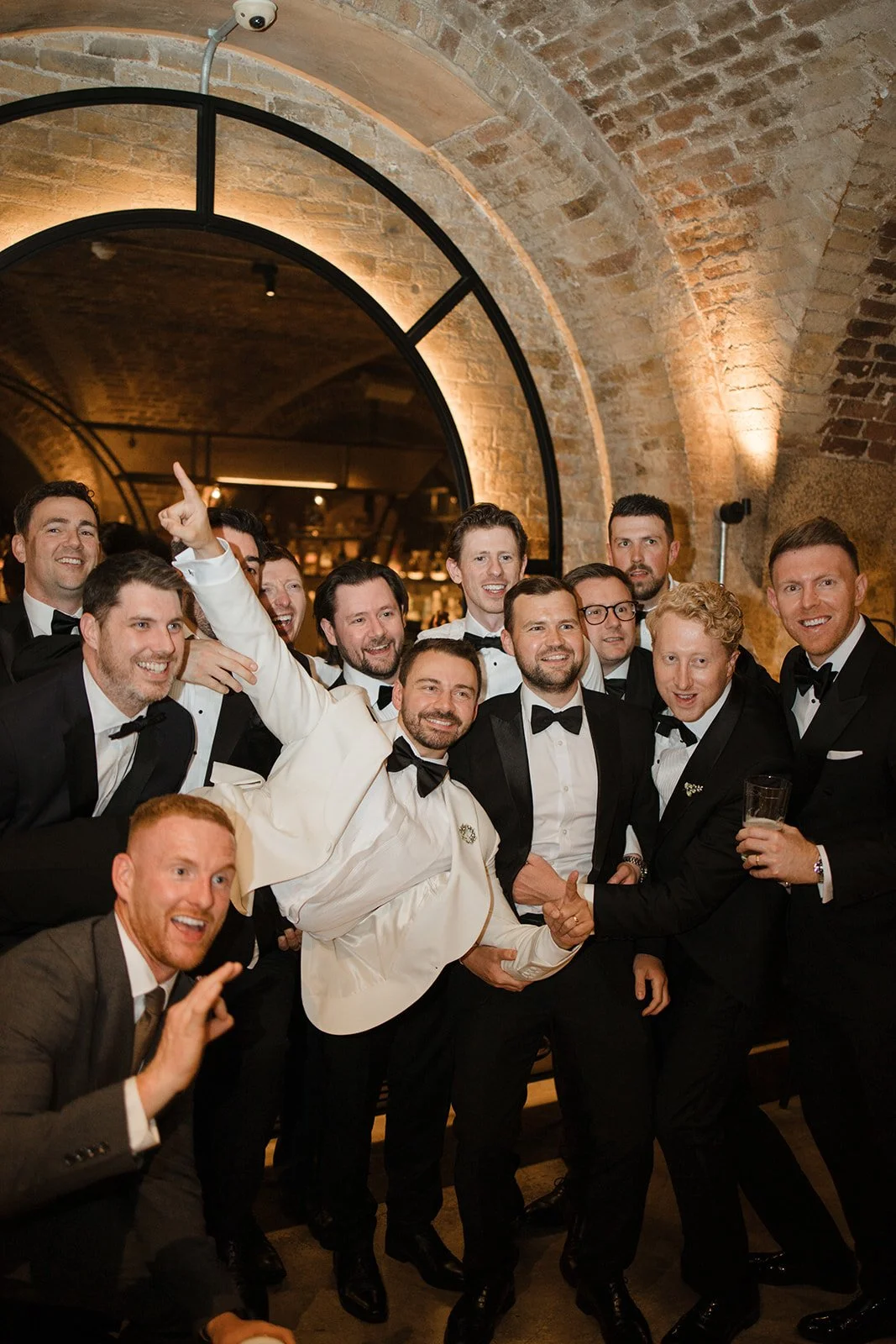A group of men in tuxedos and one man in a white suit celebrating at a party in an indoor setting with brick arch ceilings and dim lighting.