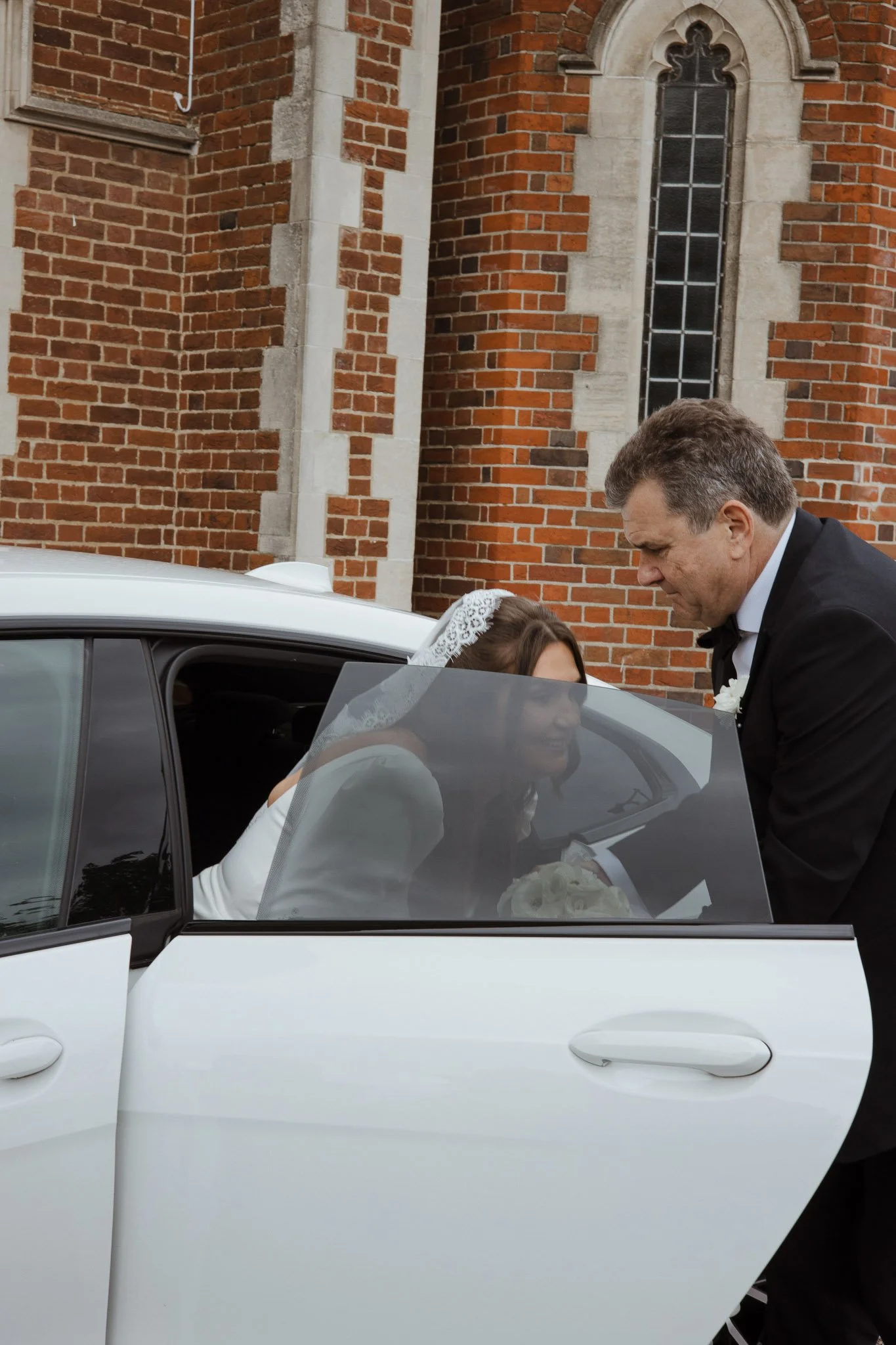A bride sitting in a white car, looking at a man in a tuxedo who is leaning in to her, with a brick church in the background.