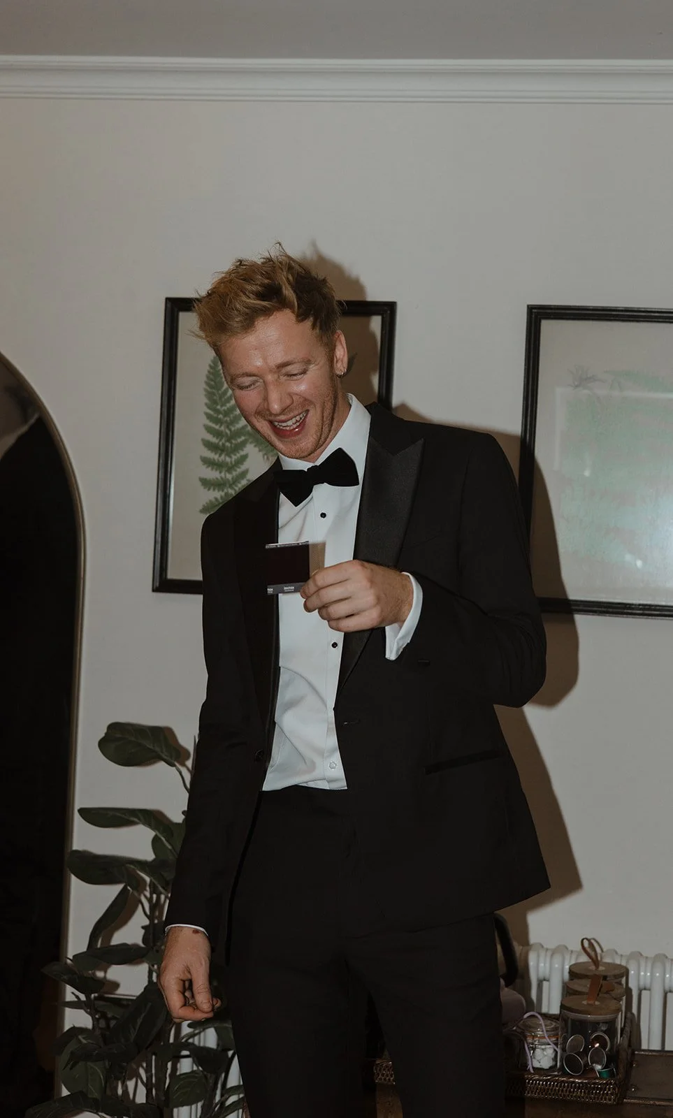 A young man in a black tuxedo and bow tie is smiling and looking at a small object in his hand, standing in a room with framed pictures and houseplants.