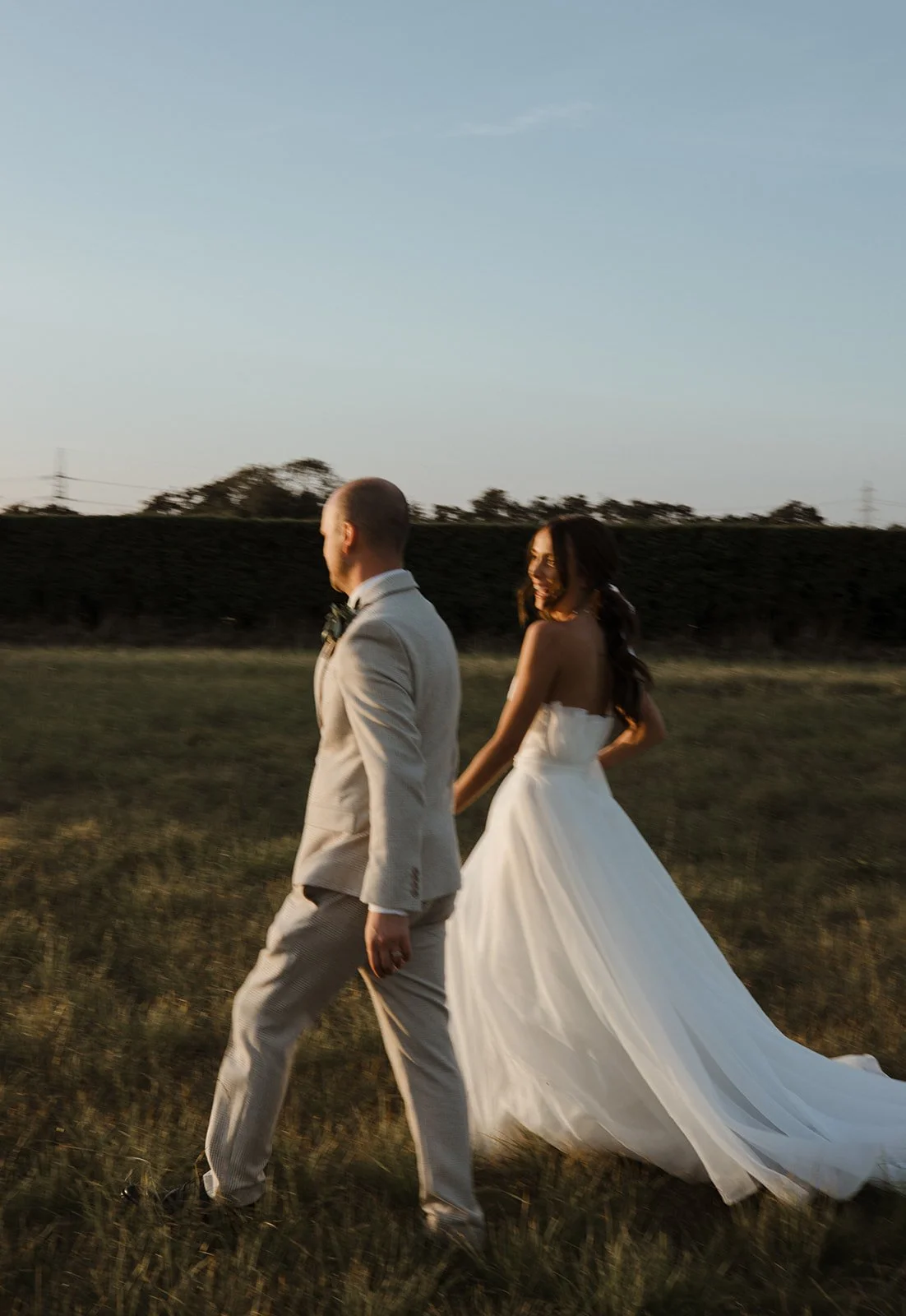 A bride and groom walking hand-in-hand in a field during sunset, with the bride smiling and wearing a white wedding gown and the groom in a light-colored suit.