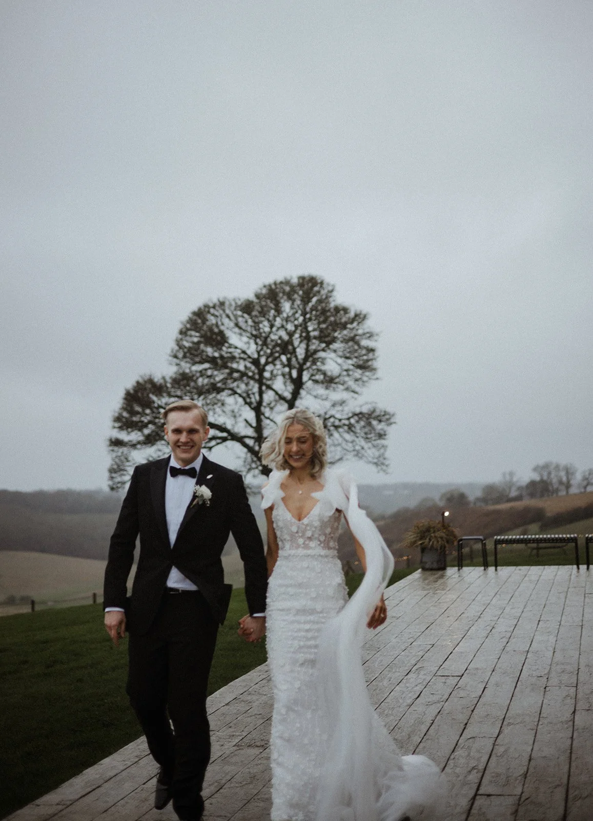 A newlywed couple walking hand in hand outdoors on a wooden deck, dressed in formal wedding attire, with a large tree and rolling hills in the background.