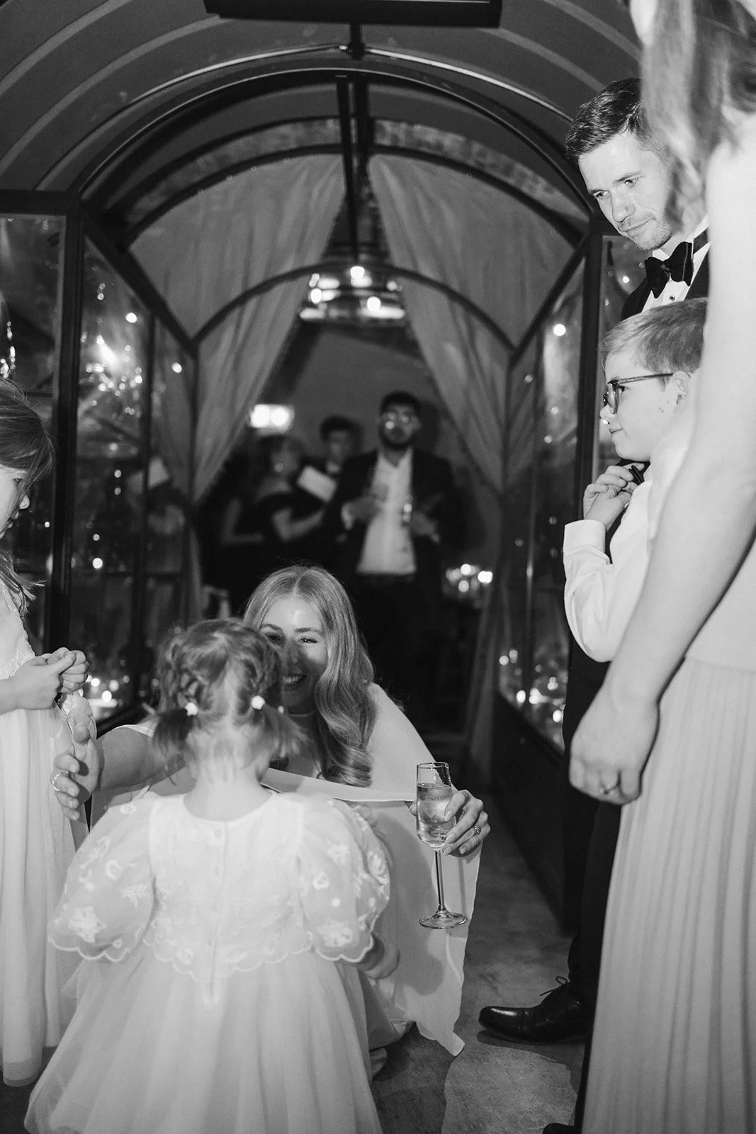 A wedding reception with guests gathered around a woman in a white dress holding a glass of champagne, engaging with children. The scene appears festive, set under a glass enclosure with reflections and string lights in the background.