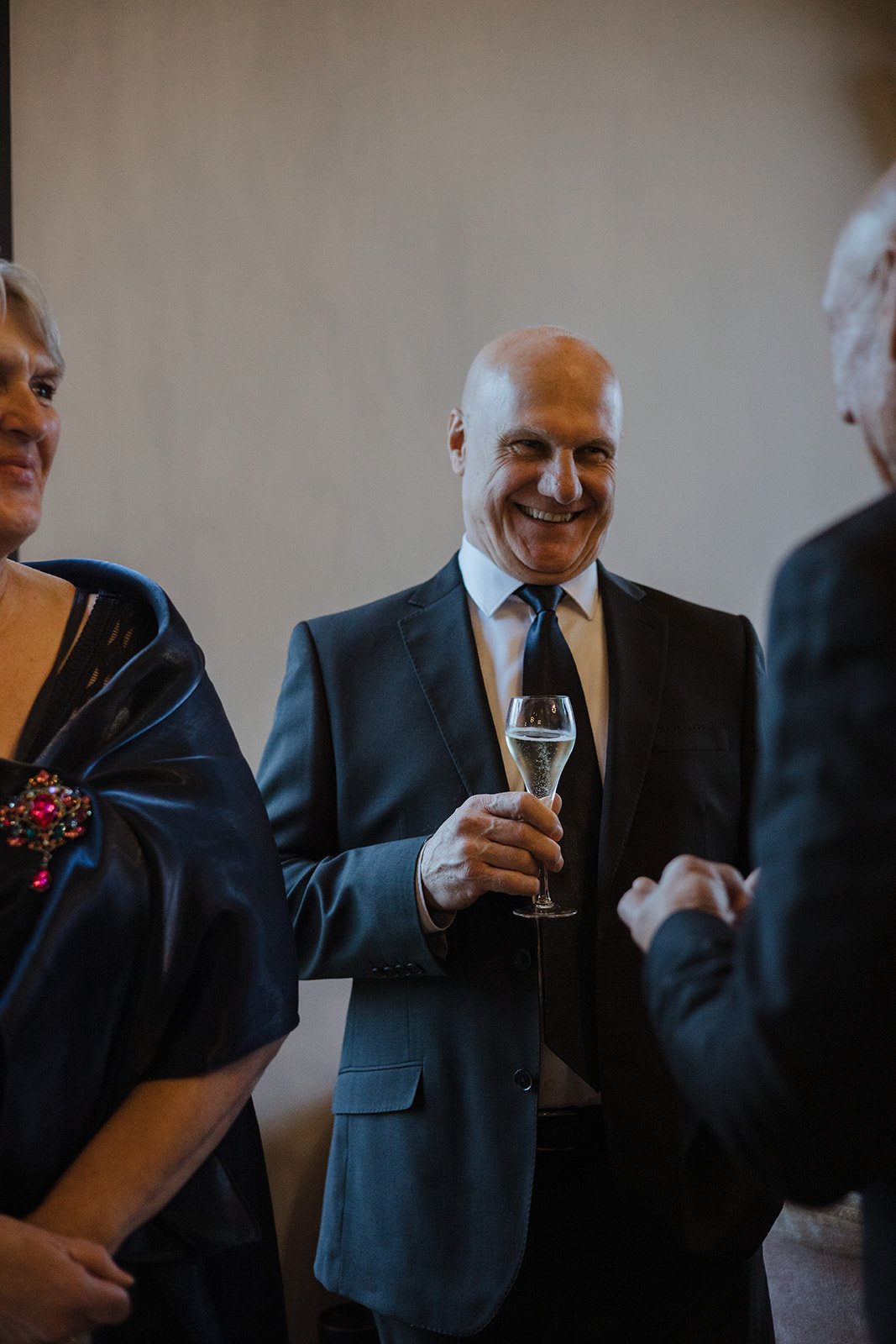 A man in a black suit and tie smiling and holding a glass of champagne at a social gathering.
