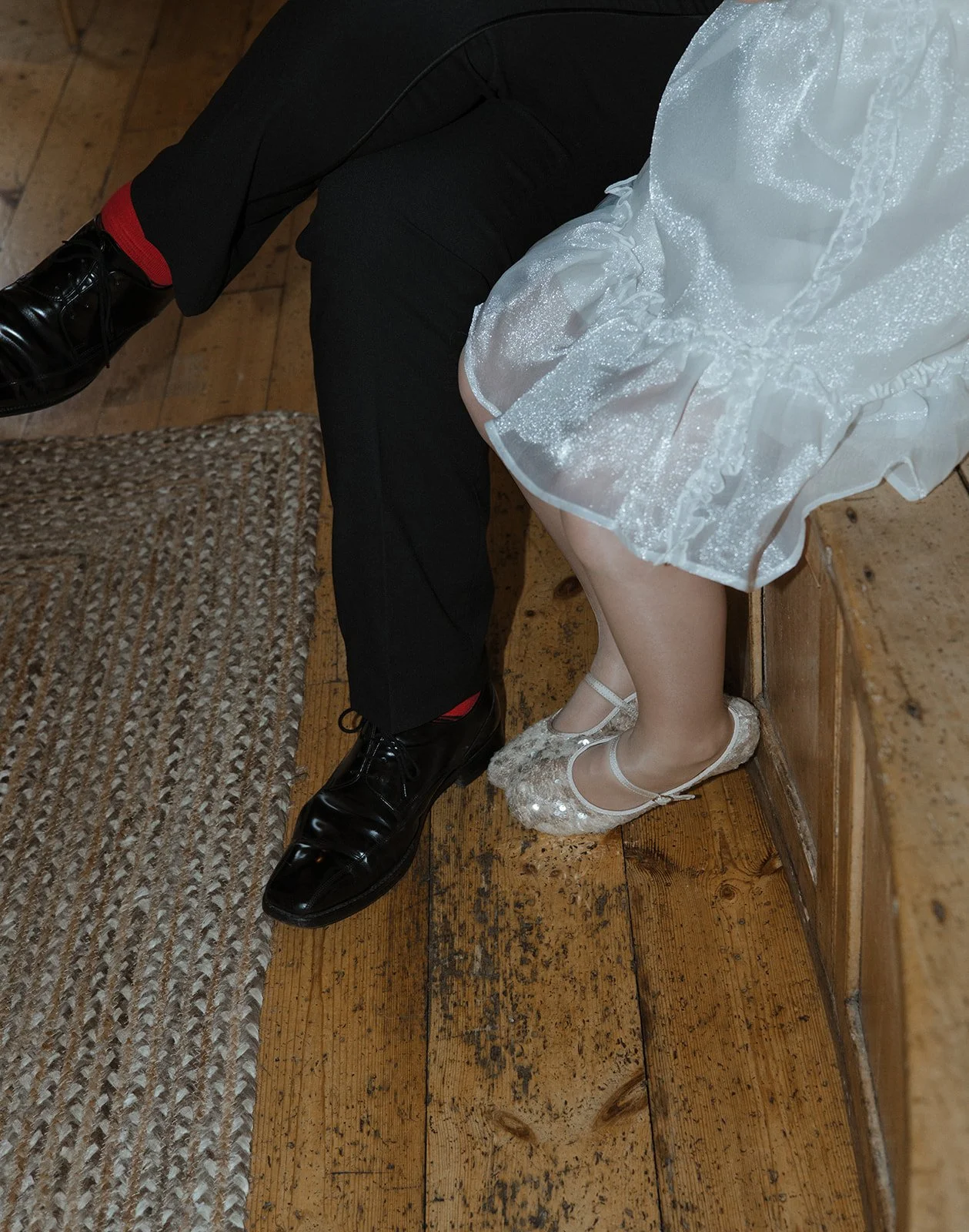 Close-up of a man and woman sitting together, showcasing their legs and shoes. The man wears black dress shoes, black pants, and red socks, while the woman wears sparkly ballet flats, with their legs crossed.