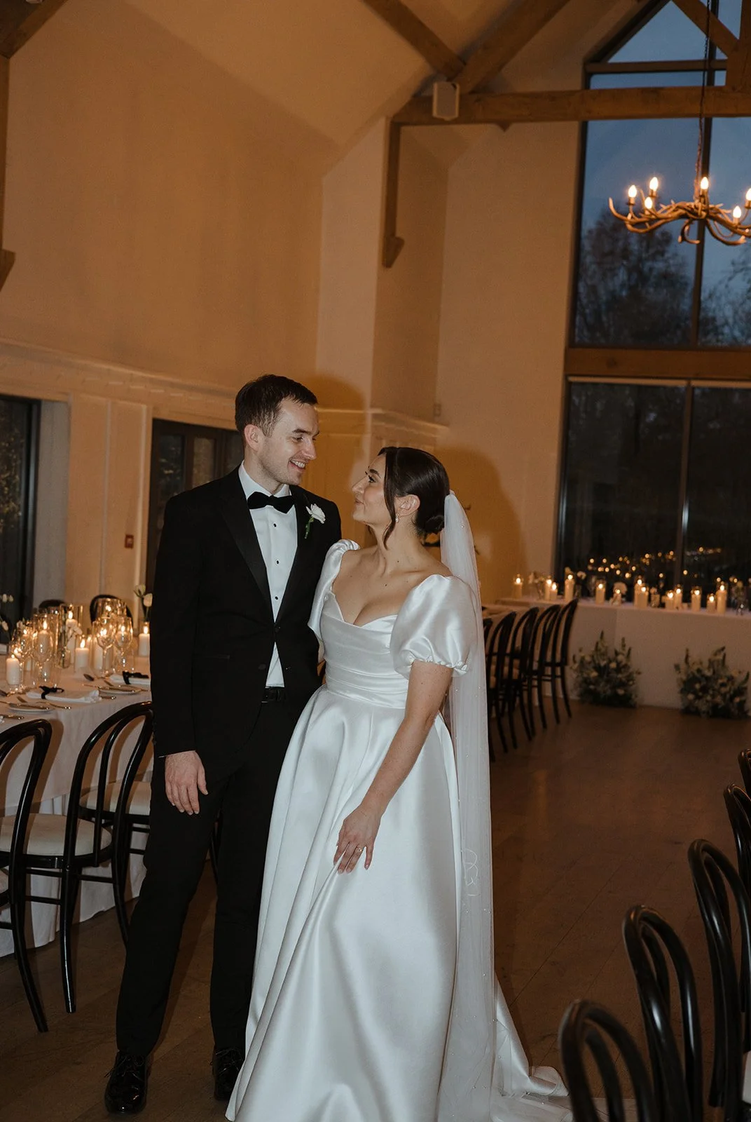 A newlywed couple dressed in wedding attire, standing close and smiling at each other inside a decorated wedding venue.