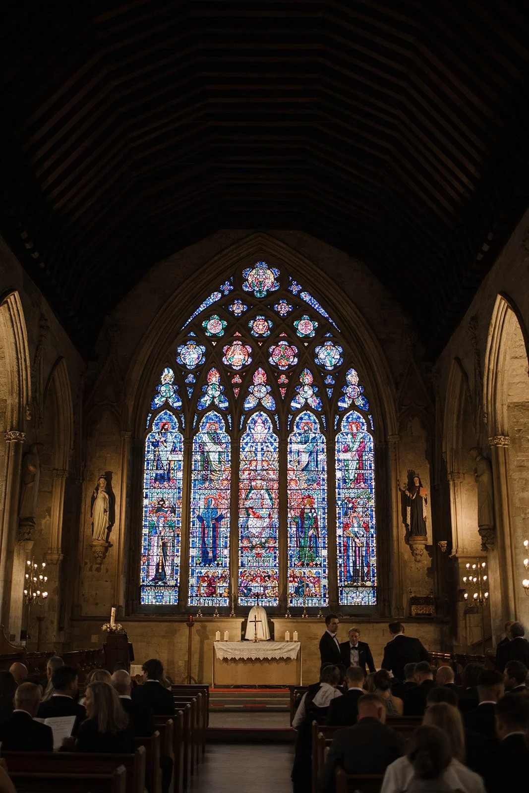 Interior of a church with stained glass windows depicting religious figures, an altar with candles, and people dressed formally attending a service or event.