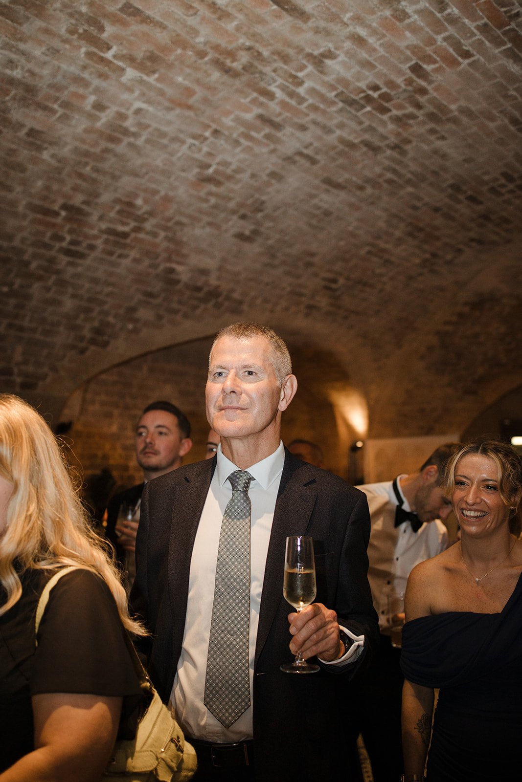 An indoor gathering with a man in a suit holding a glass of champagne, surrounded by smiling people under a brick ceiling.