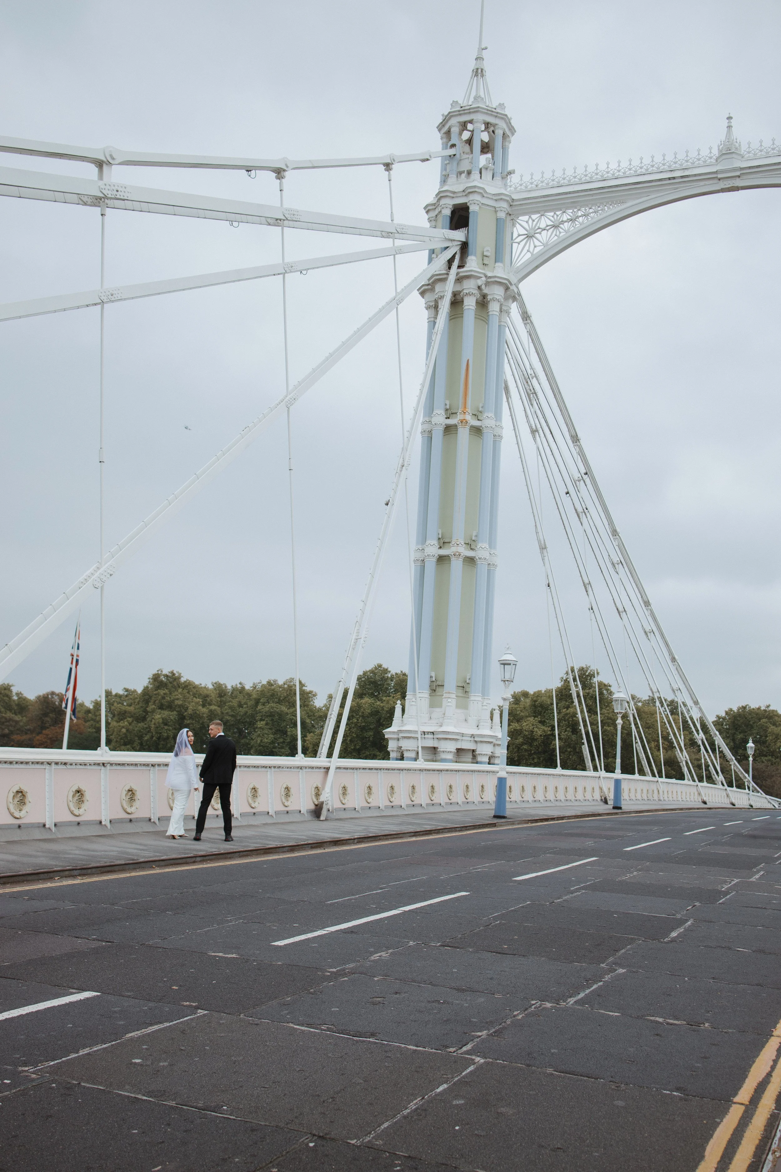 A bride and groom stand facing each other on a bridge with a front view. Behind them is a large white ferris wheel with a decorative top. The sky is cloudy, and there are trees in the background.