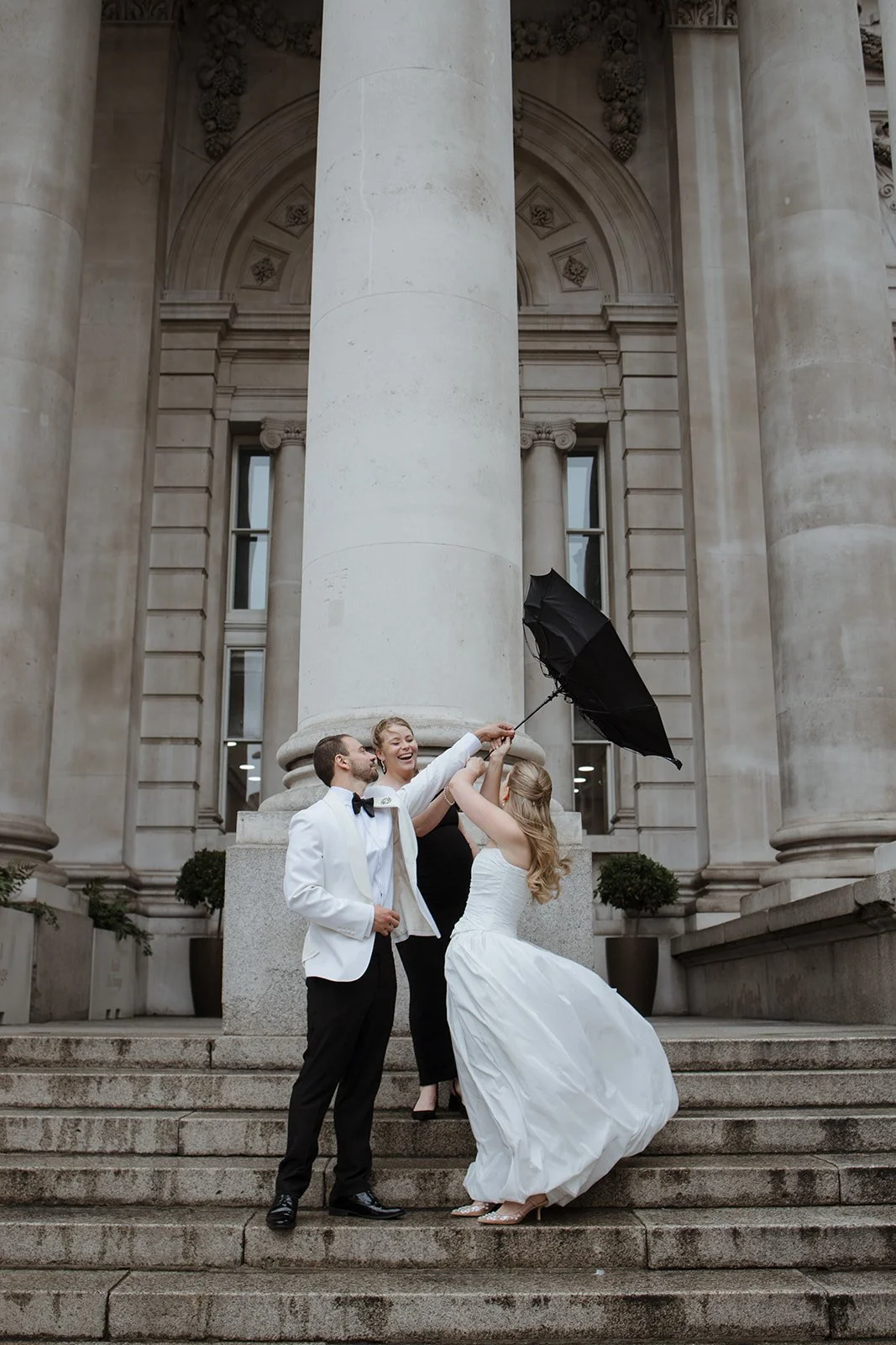 A bride in a white wedding gown, a groom in a white tuxedo jacket with a black bow tie, and a woman in black celebrate on the steps of a grand stone building. The bride is holding a black umbrella that the others are lifting together, smiling and lau