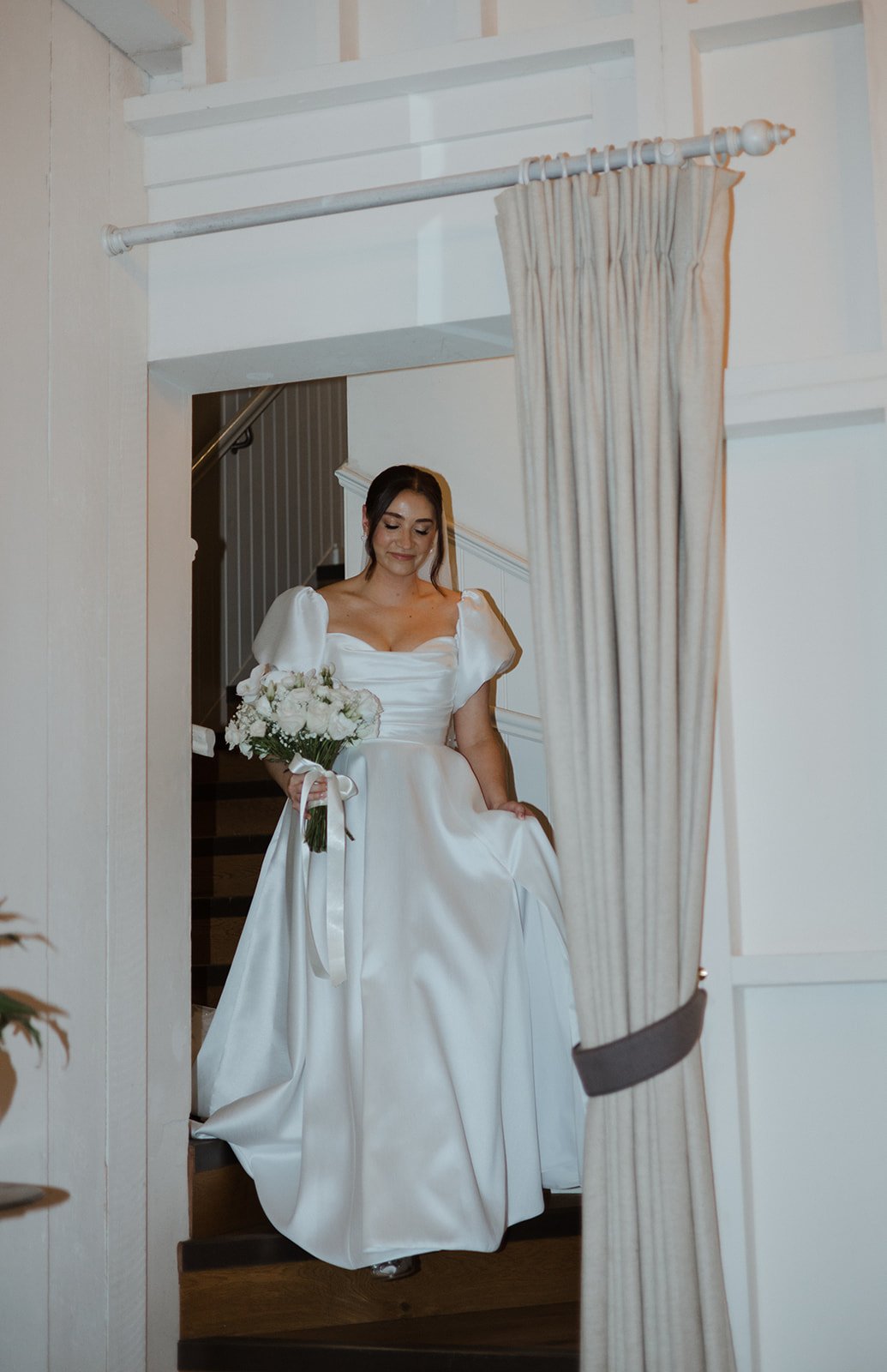 A bride in a white wedding dress holding a bouquet of white flowers, standing on a staircase.