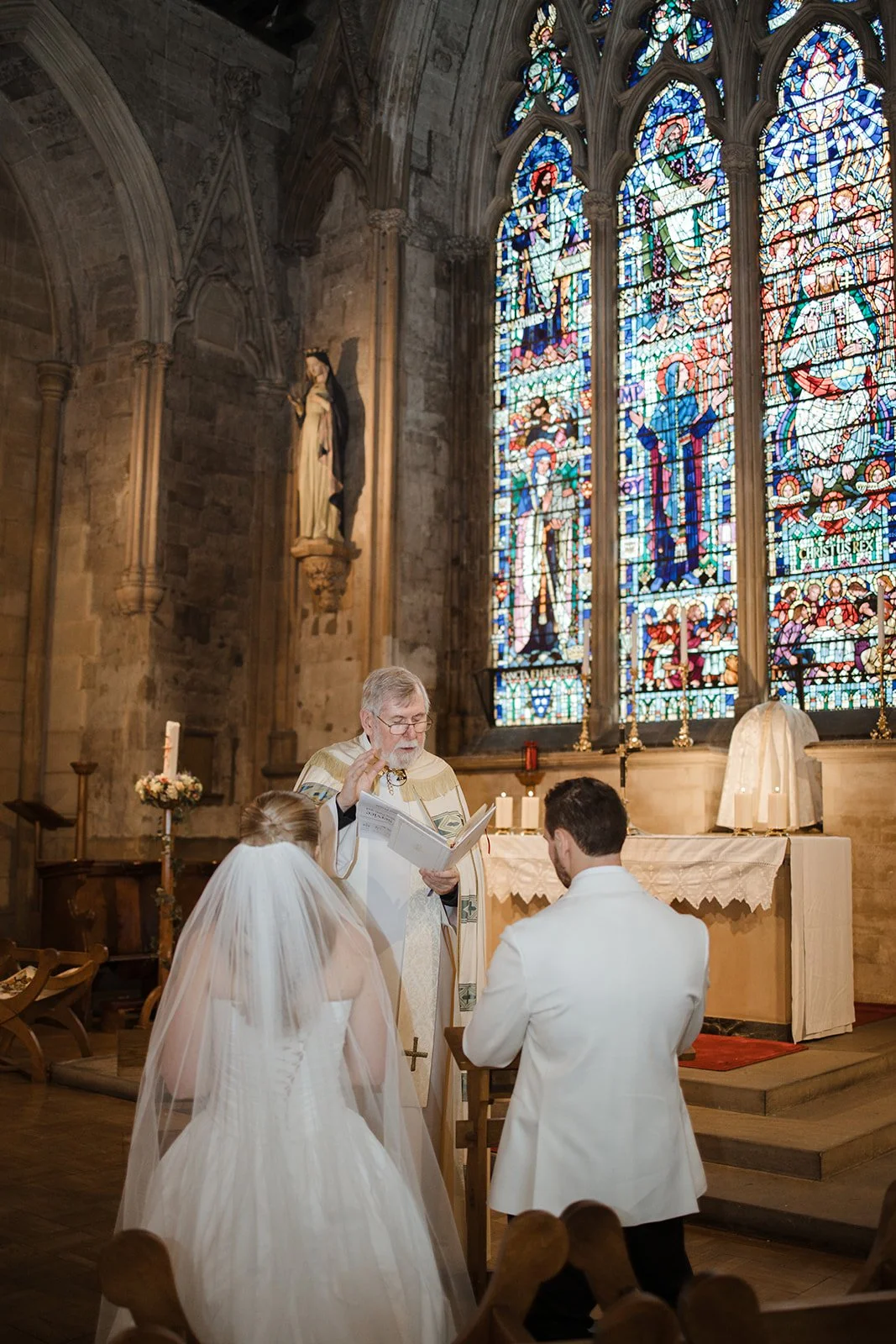 A wedding ceremony inside a church with stained glass windows. The bride and groom kneel before a priest, who is conducting the service. The bride wears a white wedding gown and veil, and the groom is in a white suit. The altar is decorated with cand