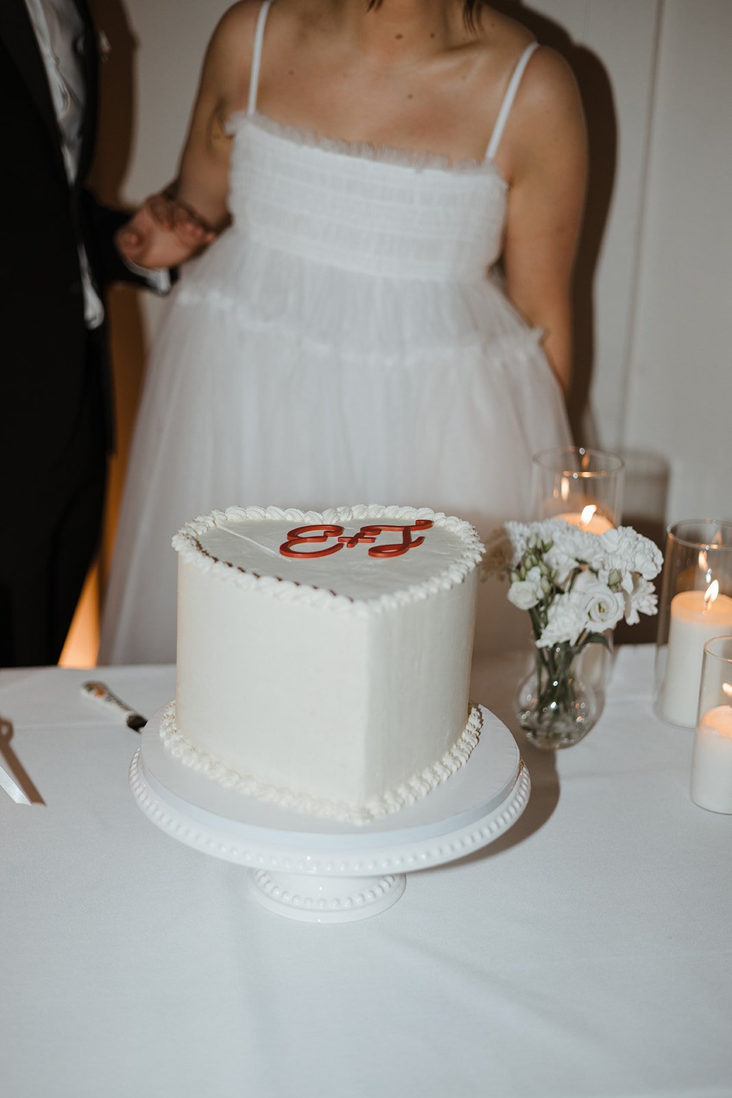 A white wedding cake with red initials "E+J" on top, placed on a white cake stand. In the background, a woman in a white dress is visible, along with white flowers and lit candles on the table.