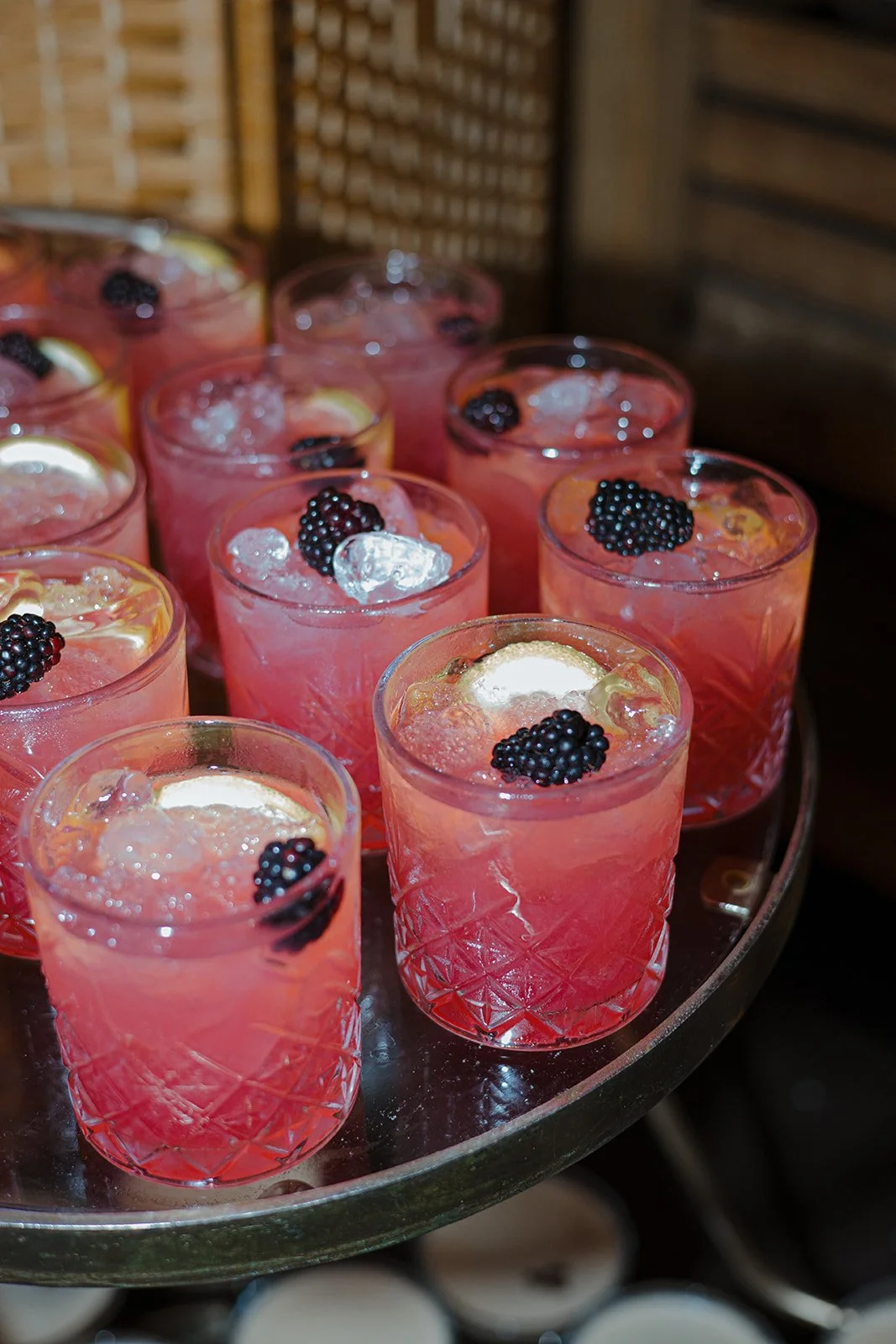 A tray of pink cocktails garnished with blackberries and lemon slices, with ice cubes, on a silver tray in a dimly lit setting.