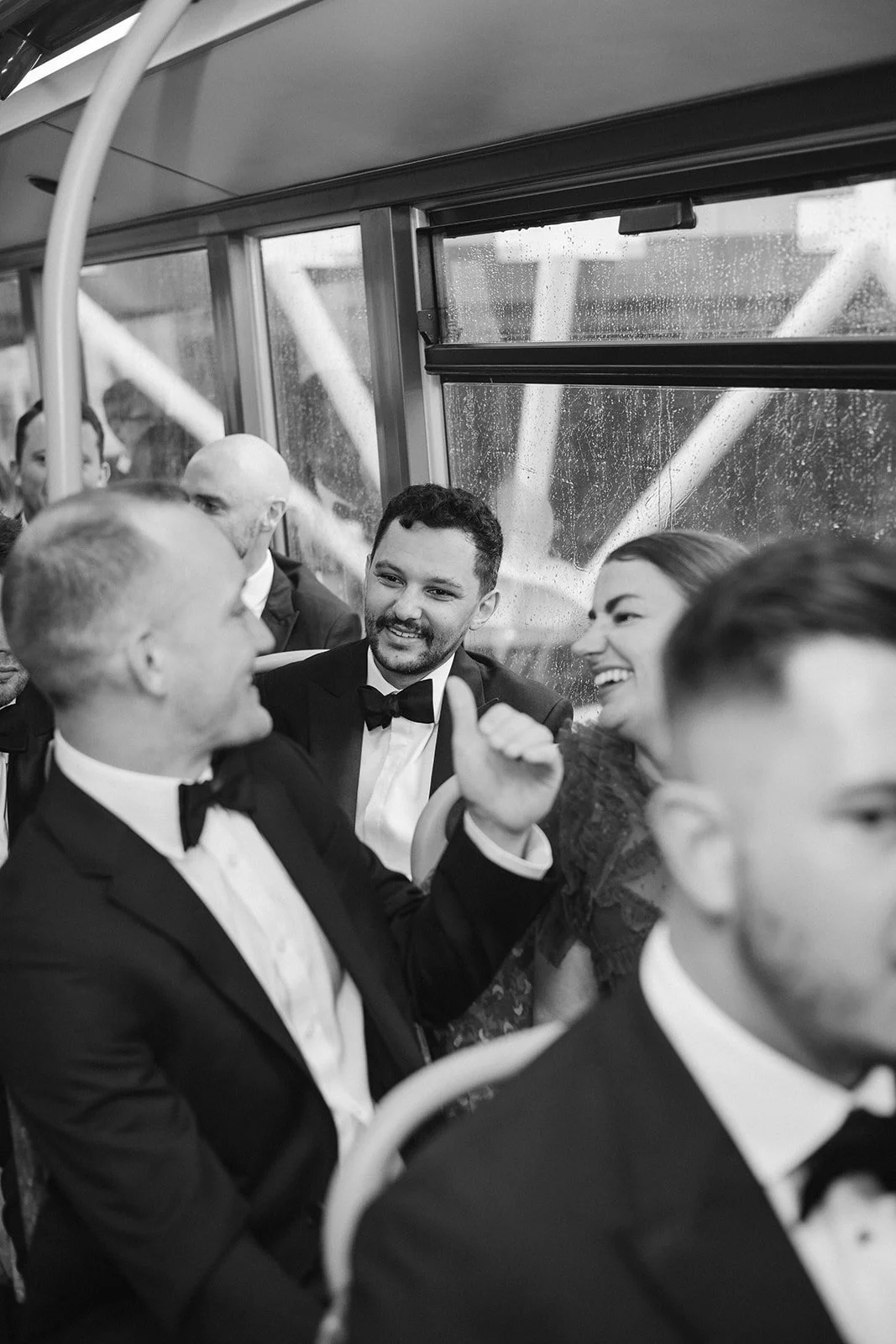 People celebrating and talking on a bus, dressed in formal tuxedos and dresses, with rainy weather outside the window.