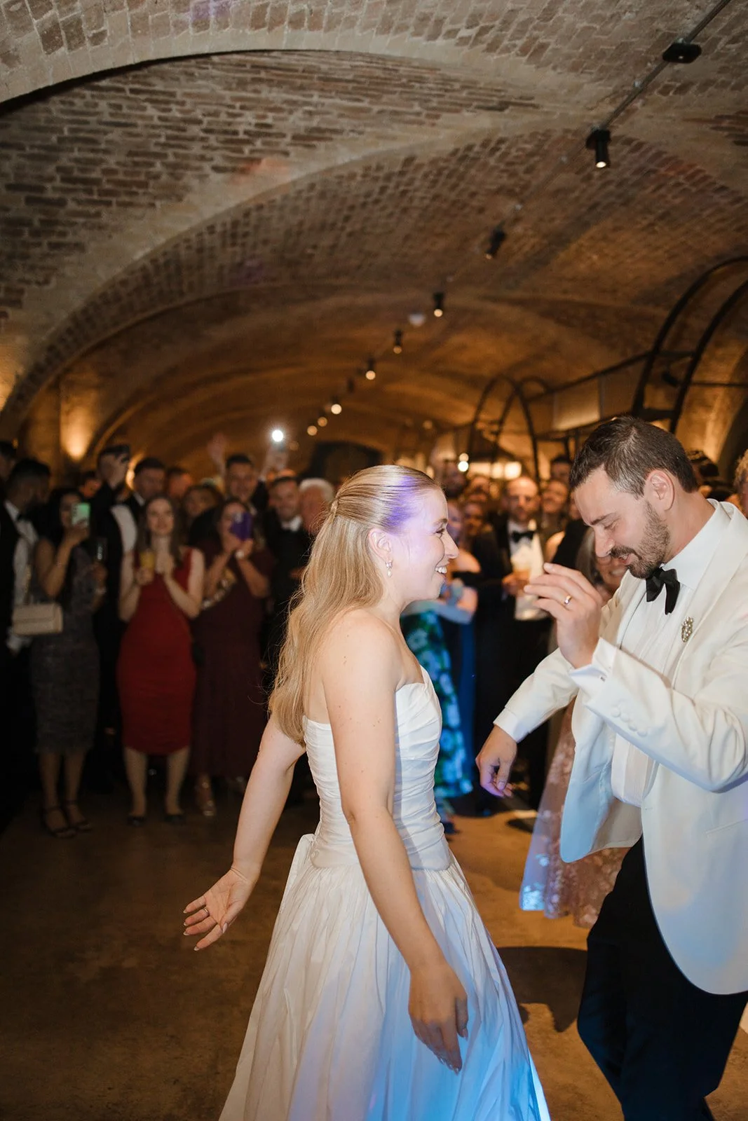 Bride and groom dancing at wedding reception with friends in background under brick vaulted ceiling.
