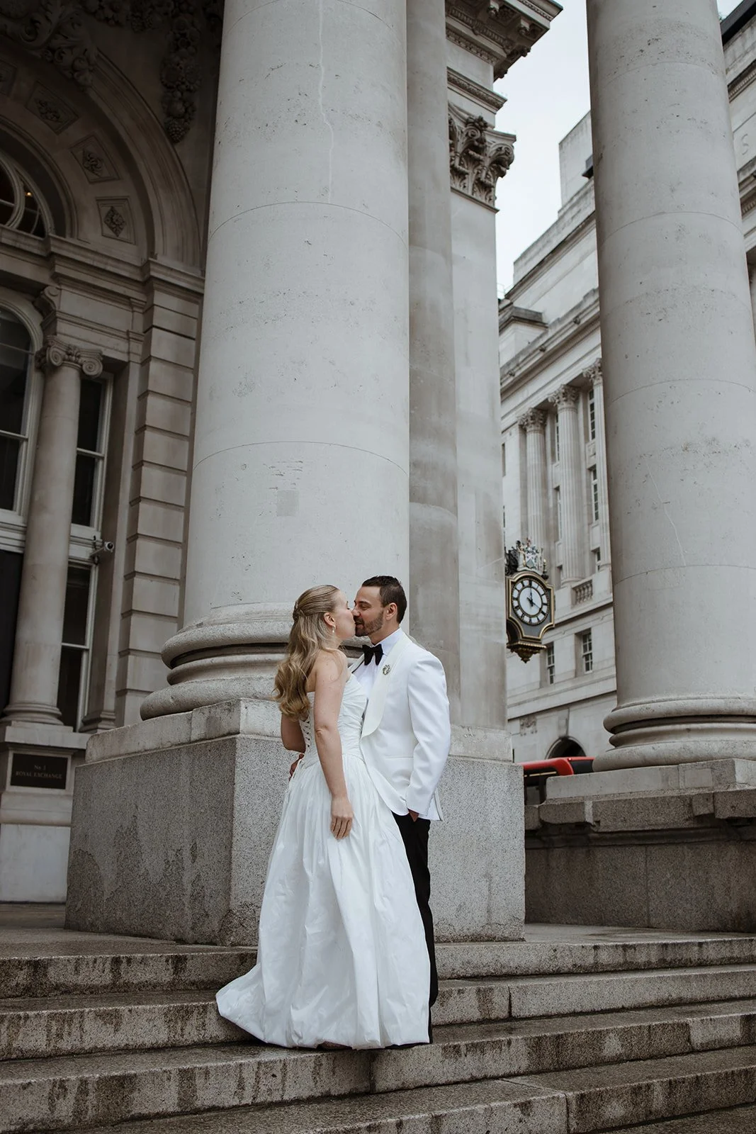 A newlywed couple, dressed in wedding attire, sharing a kiss on steps outside a historic building with large stone columns and a clock in the background.